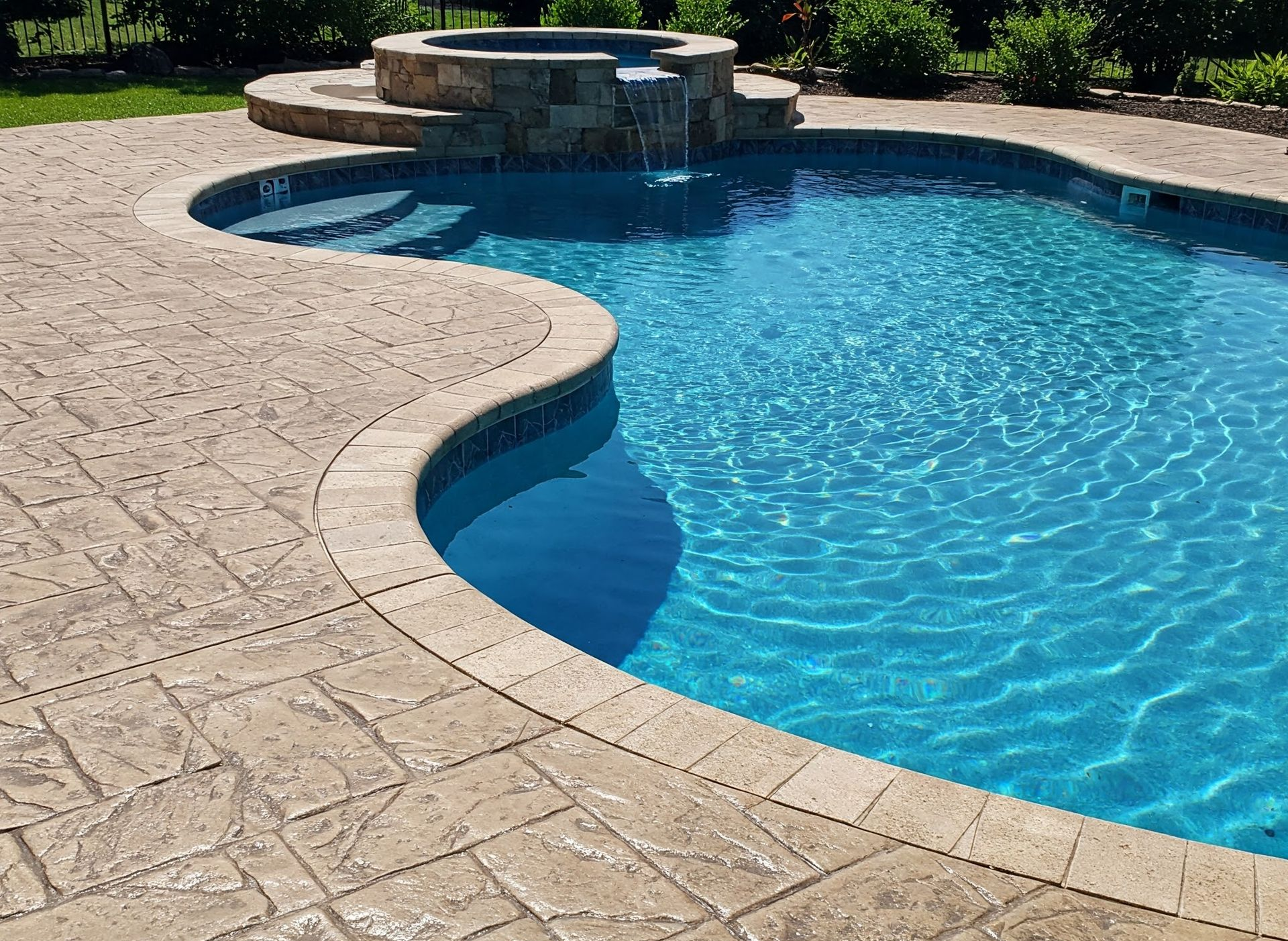 Pool with blue water, surrounded by textured stamped concrete. A small waterfall is in the background.
