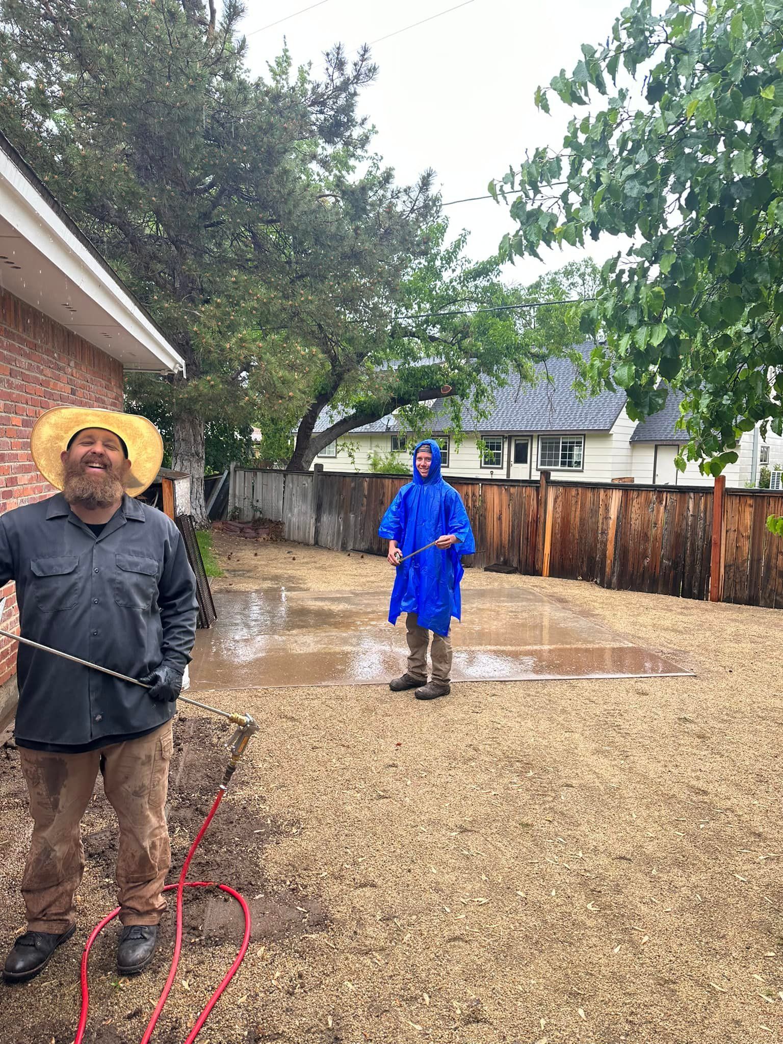 A man in a blue raincoat is spraying water on a gravel driveway.