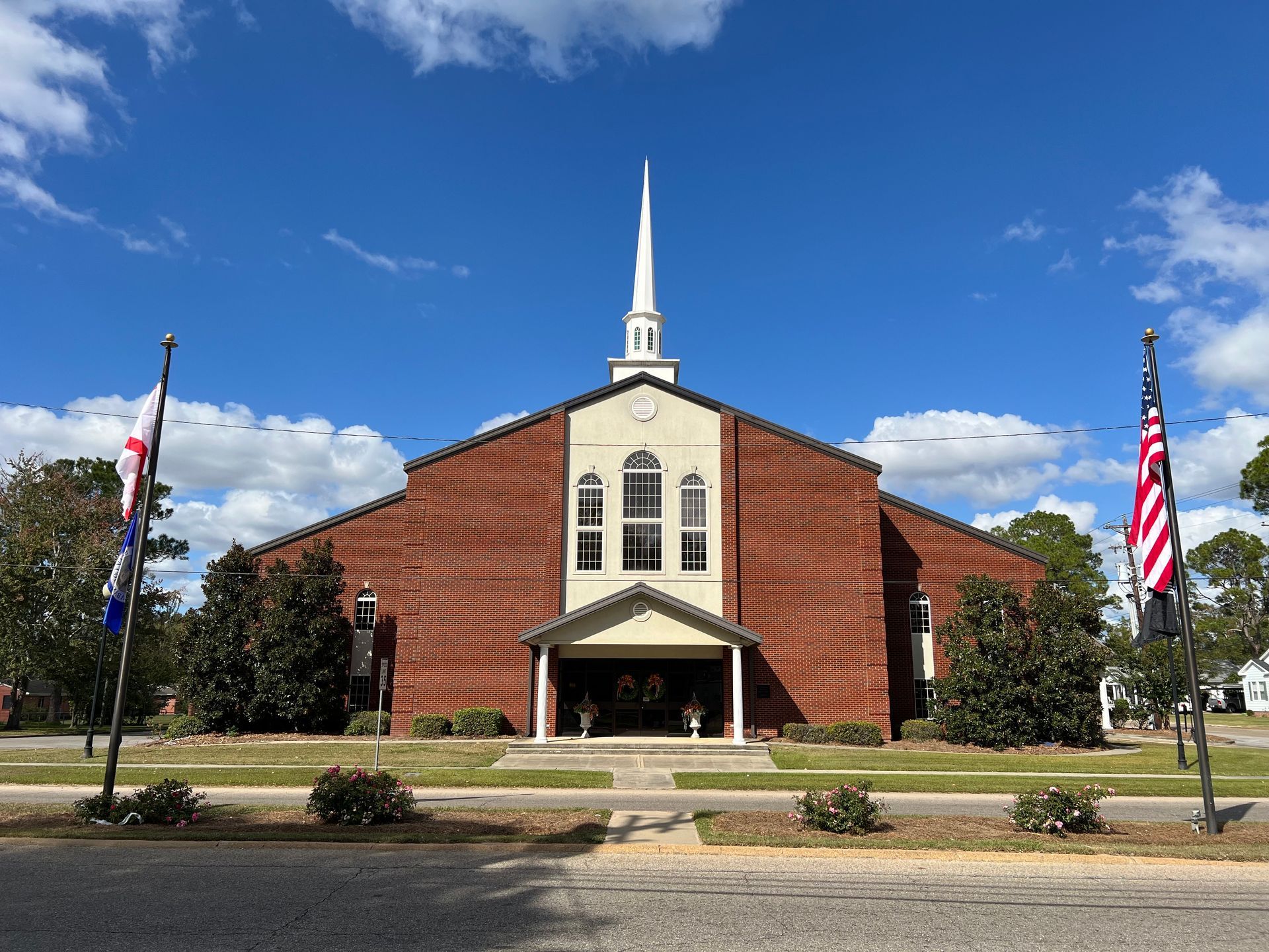 A First Baptist Church with a steeple and two american flags in front of it.