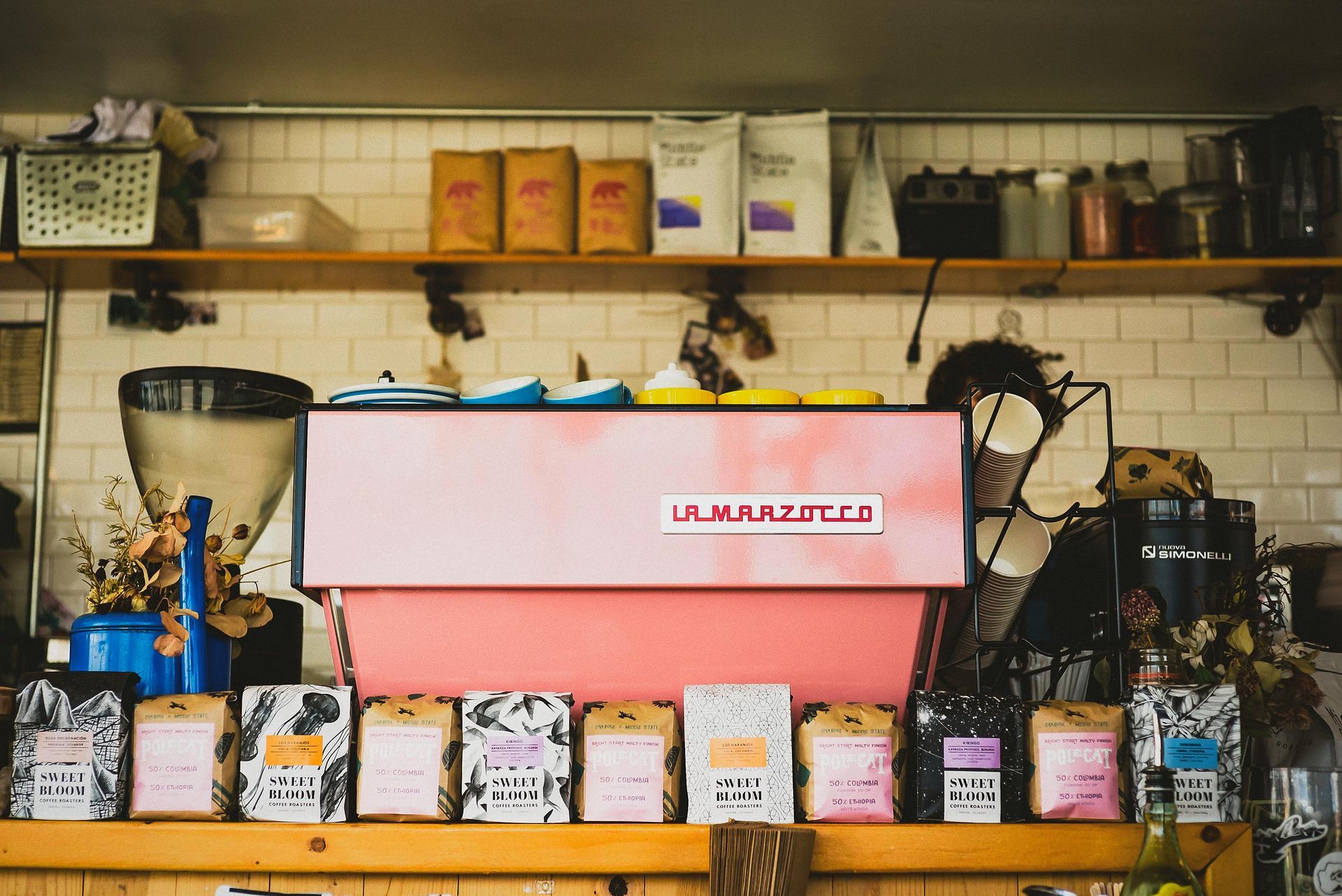 Pink espresso machine behind counter with coffee bean packages, shelves of supplies in a coffee shop.