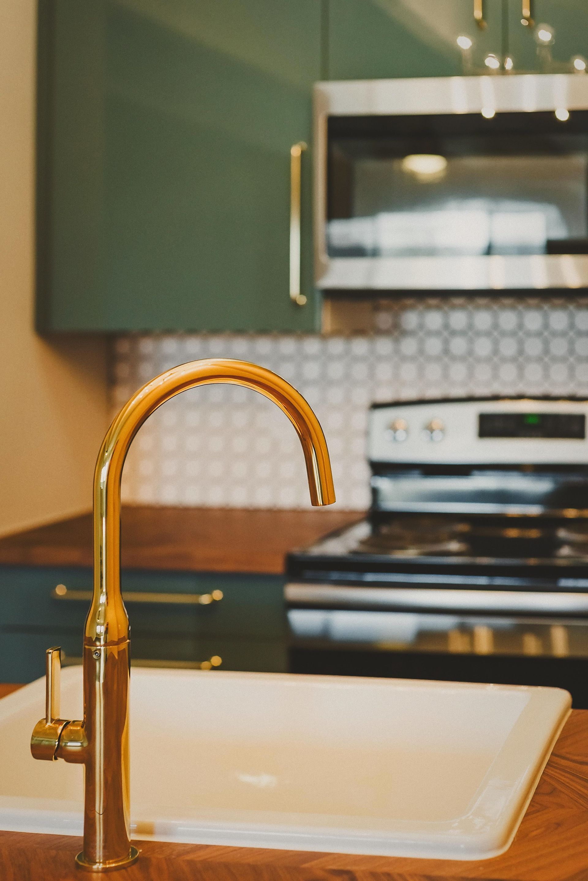 Gold kitchen faucet over a white sink, with green cabinets and a black stove in the background.