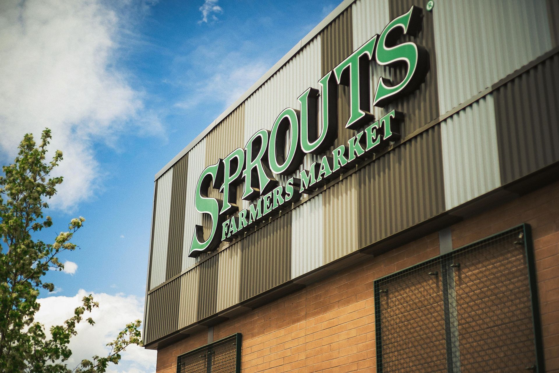 Sprouts Farmers Market sign on a building exterior, green lettering against a metallic facade, cloudy sky.
