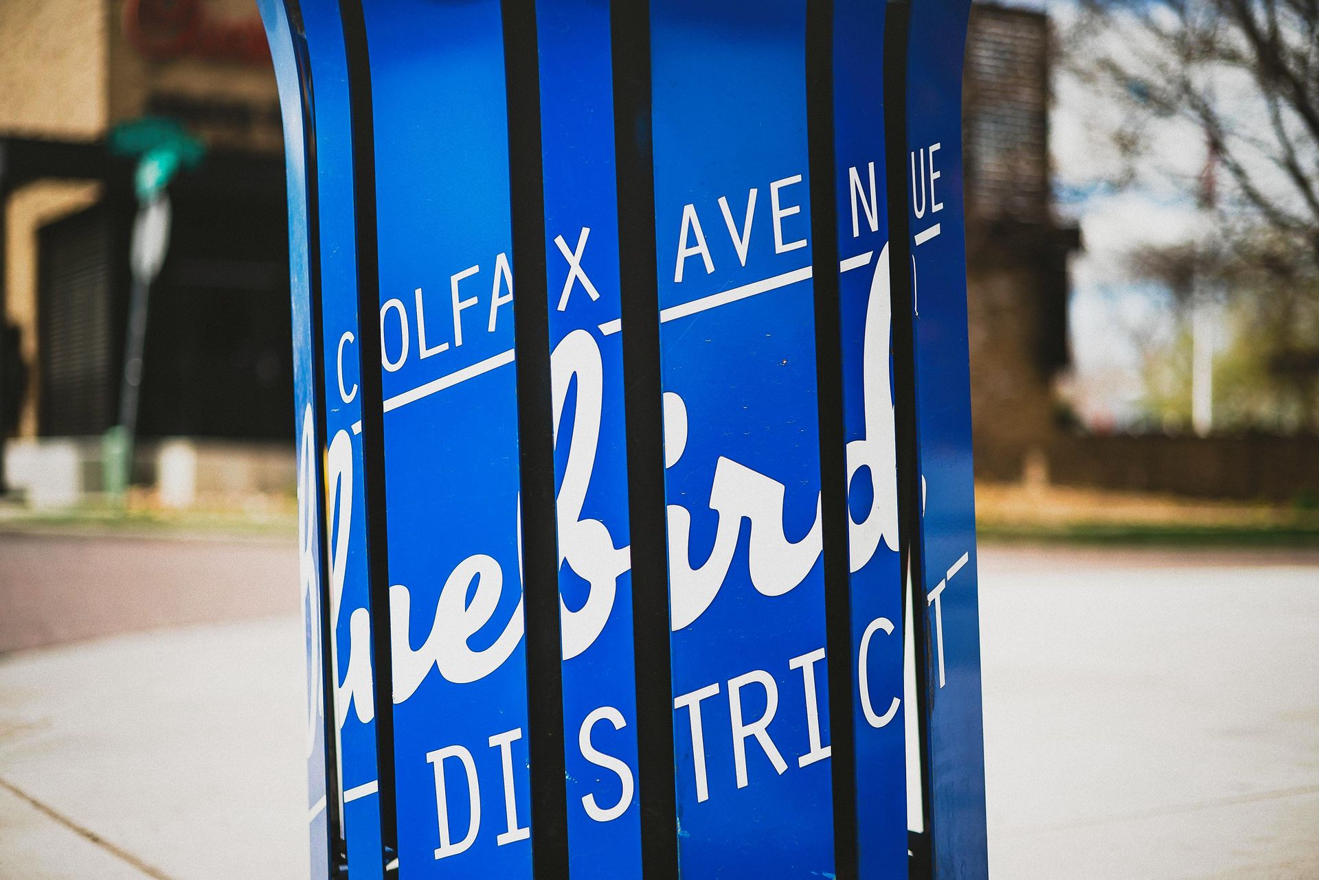 Sign for Colfax Avenue's Bluebird District; blue background, white text, obscured by black vertical lines.