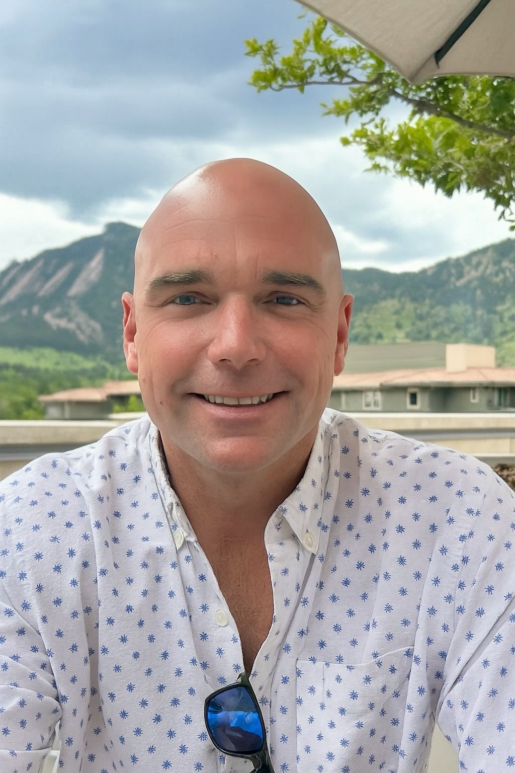 Man smiles at the camera outside. Blue and white patterned shirt. Mountain background.
