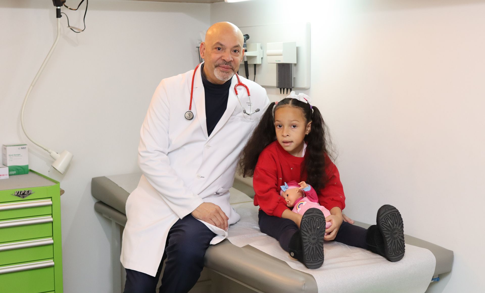 A male pediatrician is sitting on a medical table with a patient.
