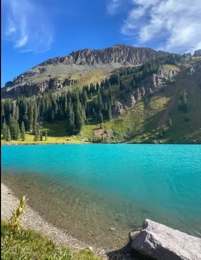 Turquoise lake at the base of a rocky mountain. Green trees and grassy areas are visible under a bright blue sky.