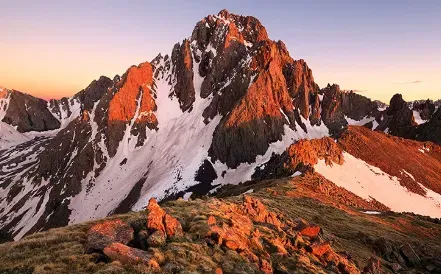 Snow-capped mountain peak bathed in golden light at sunset. Rugged terrain in foreground.