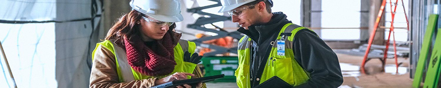 A man and a woman are standing next to each other on a construction site looking at a tablet.