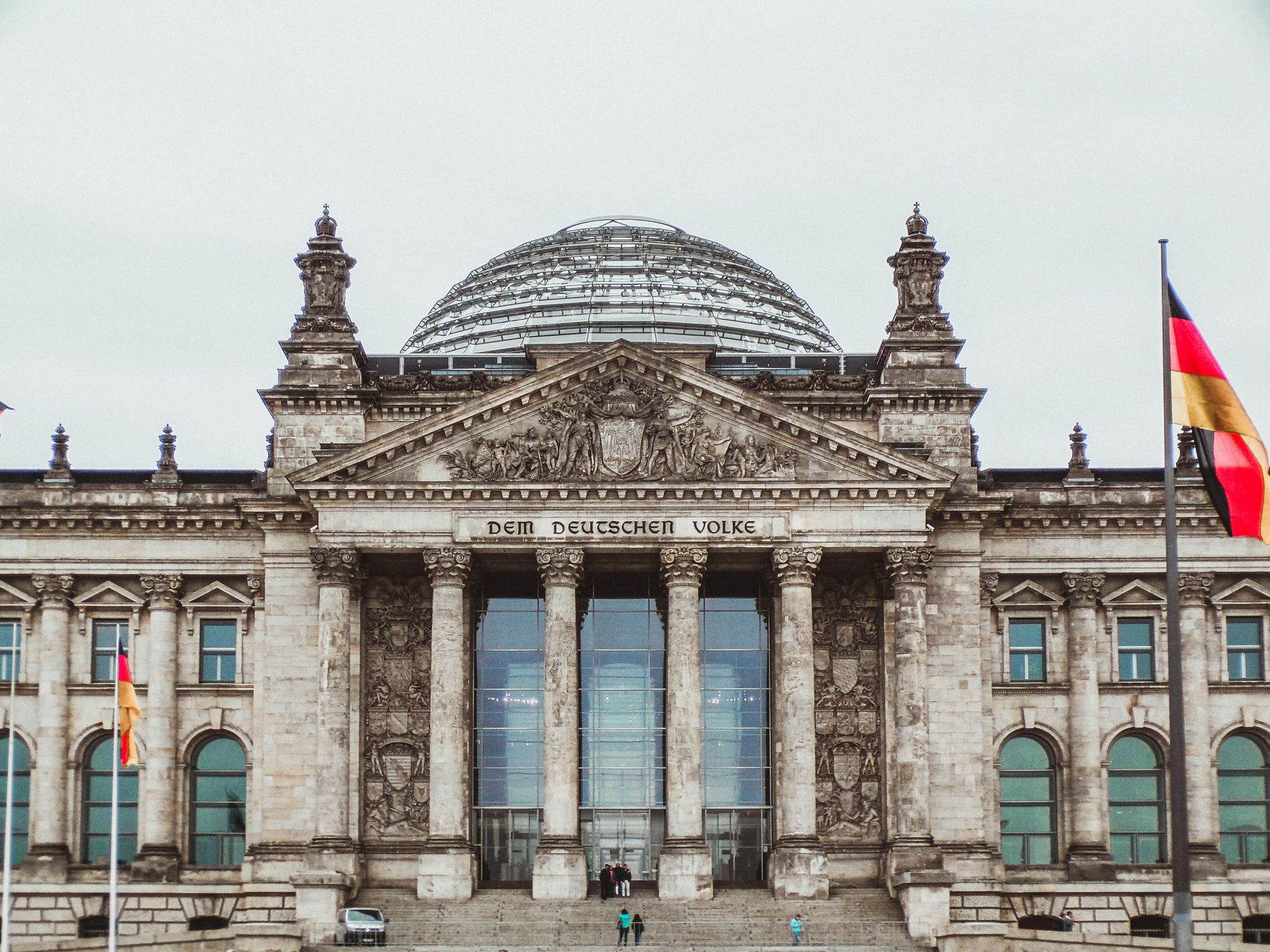 Reichstag Building in Berlin, Germany, with a glass dome and German flags on a cloudy day.
