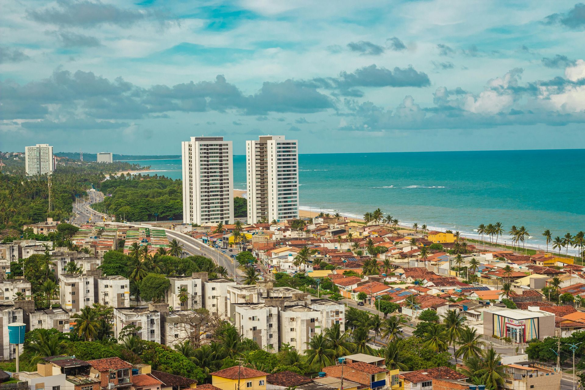 Cityscape with tall white buildings, beach, and ocean under a cloudy sky.