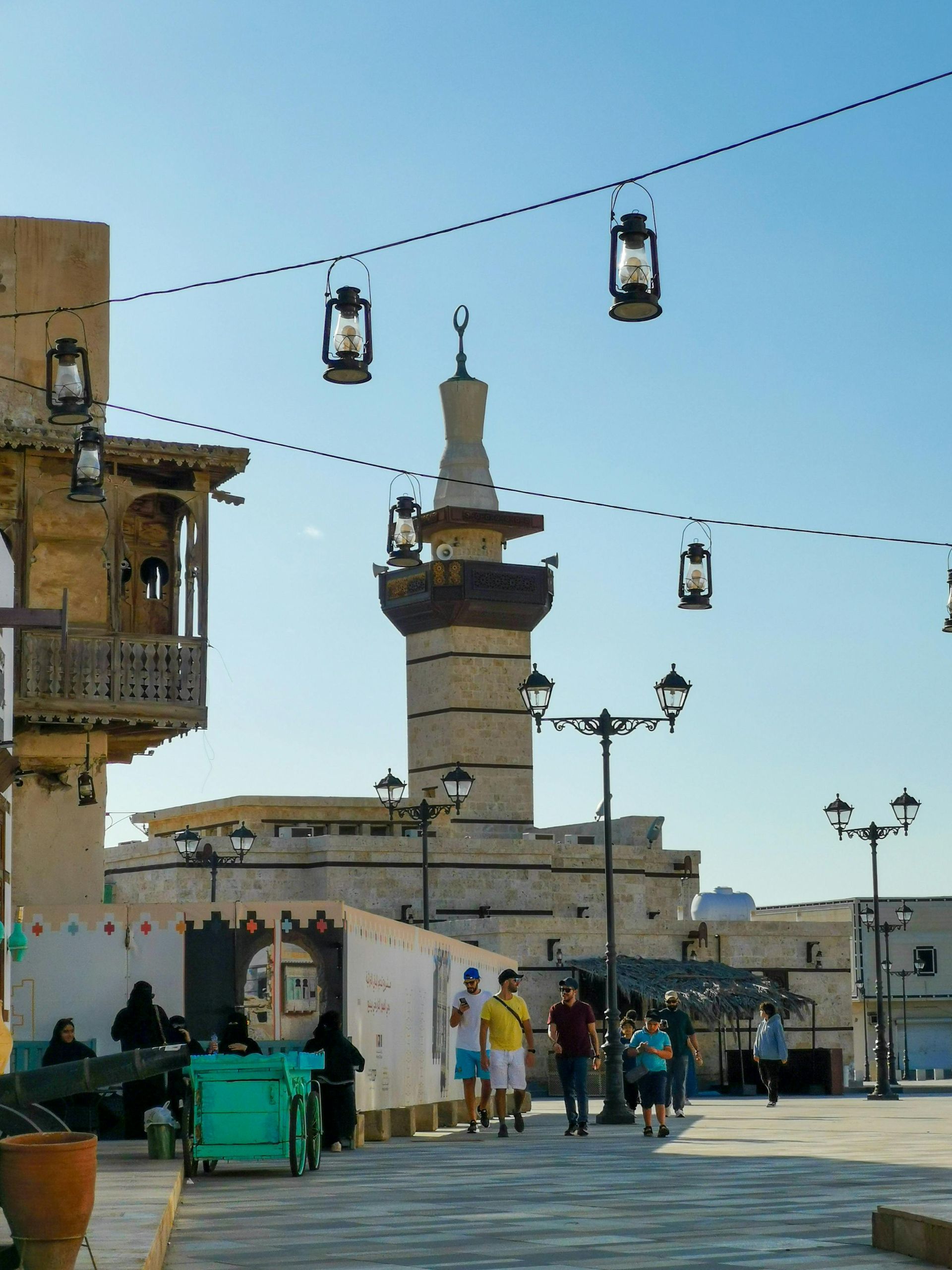 Street scene with a mosque tower, lanterns, and people walking on a sunny day.
