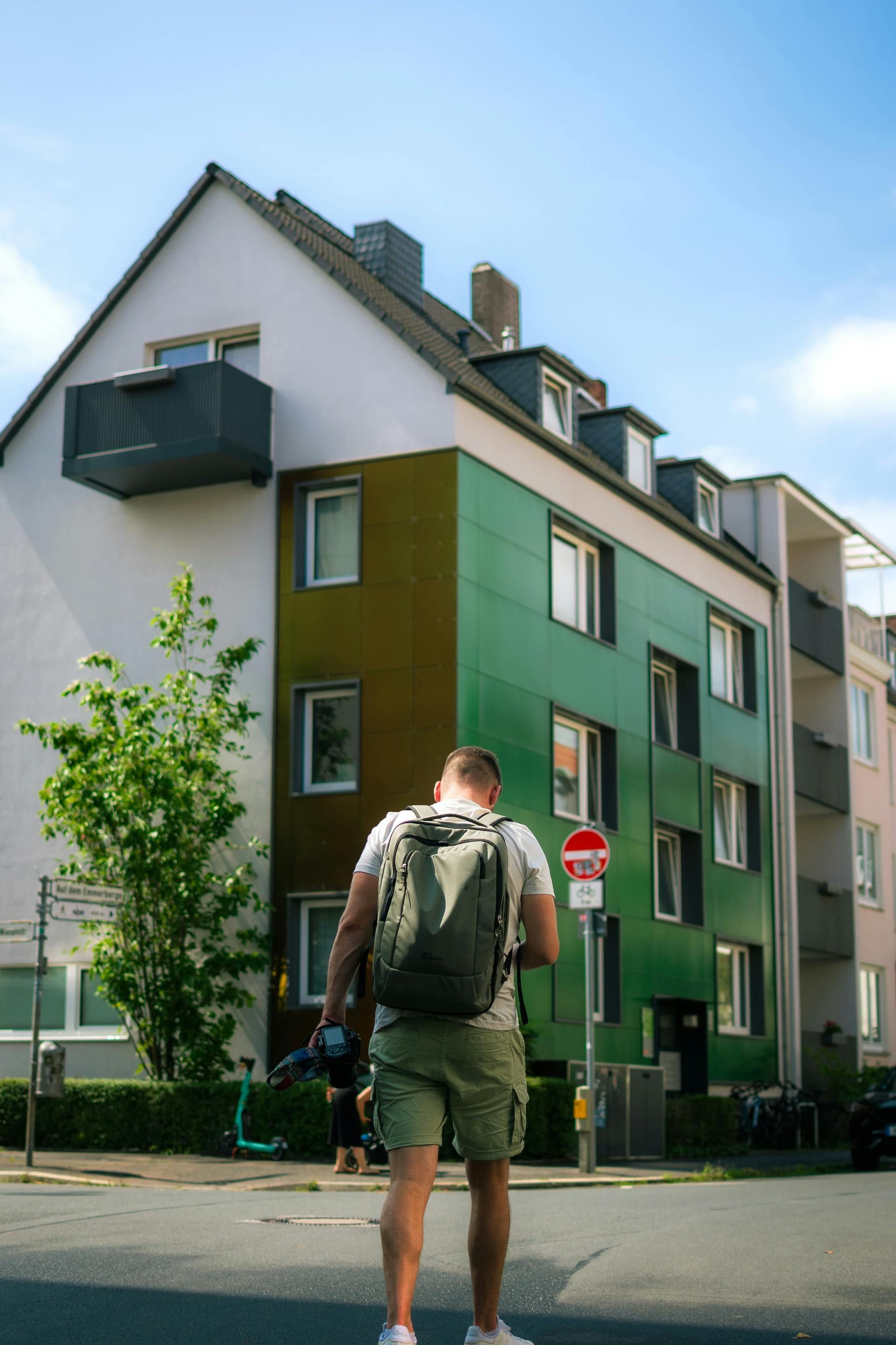 Man with backpack stands in front of colorful apartment building on a sunny day.