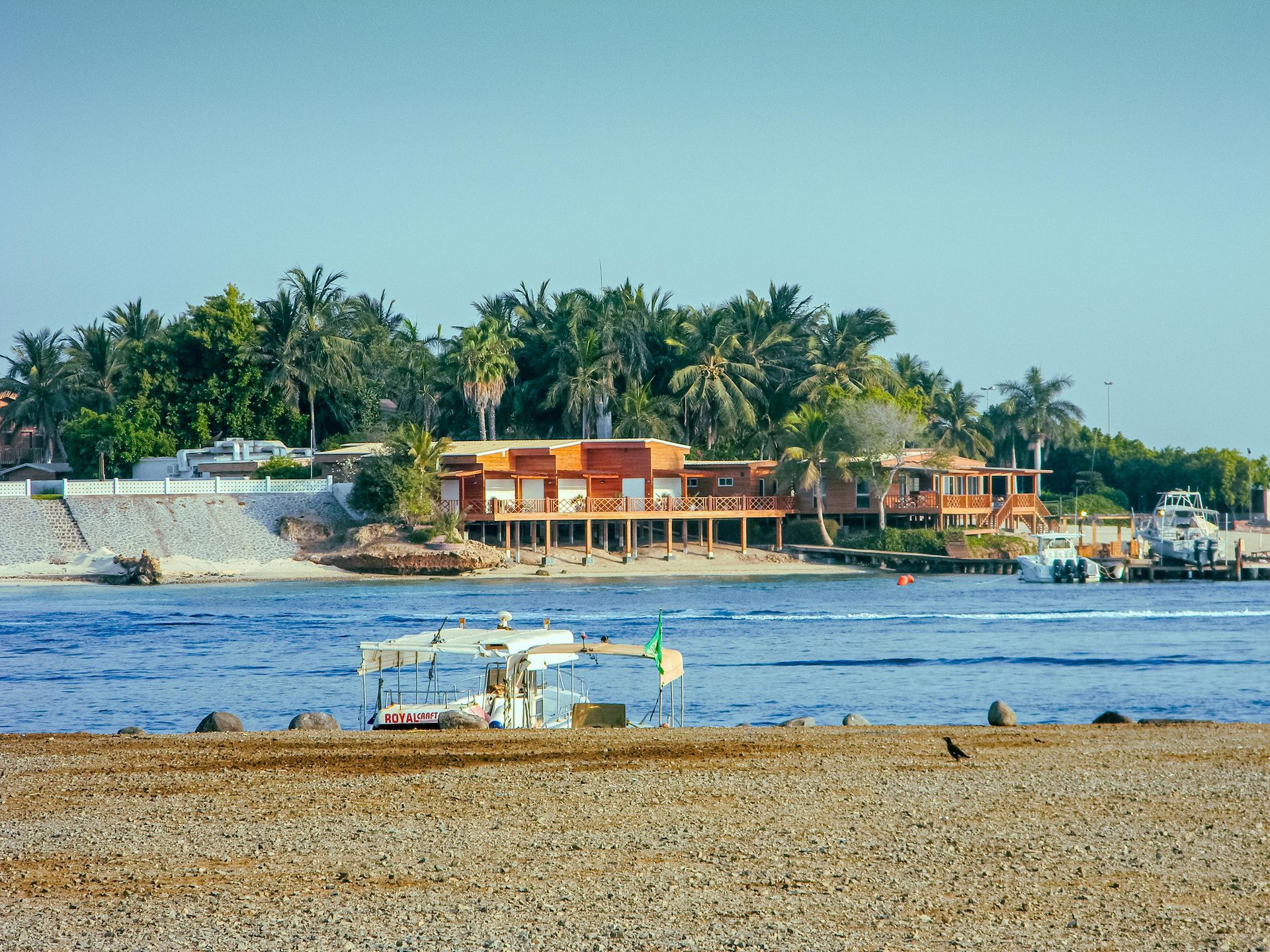 Beach scene with a red building and boats on the water; tropical trees in the background.
