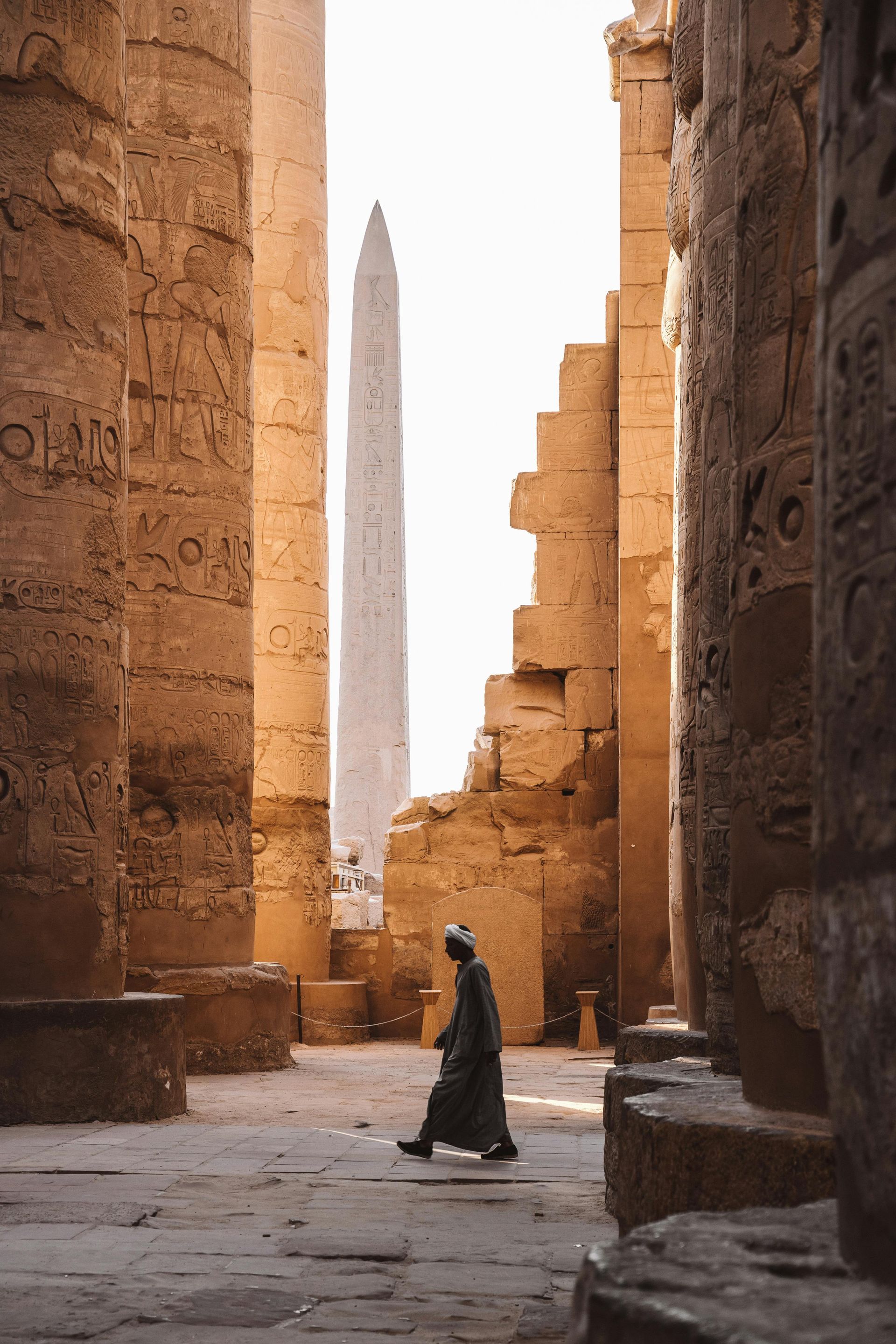 Man in traditional robes walks through ancient temple ruins toward a tall obelisk in Egypt.