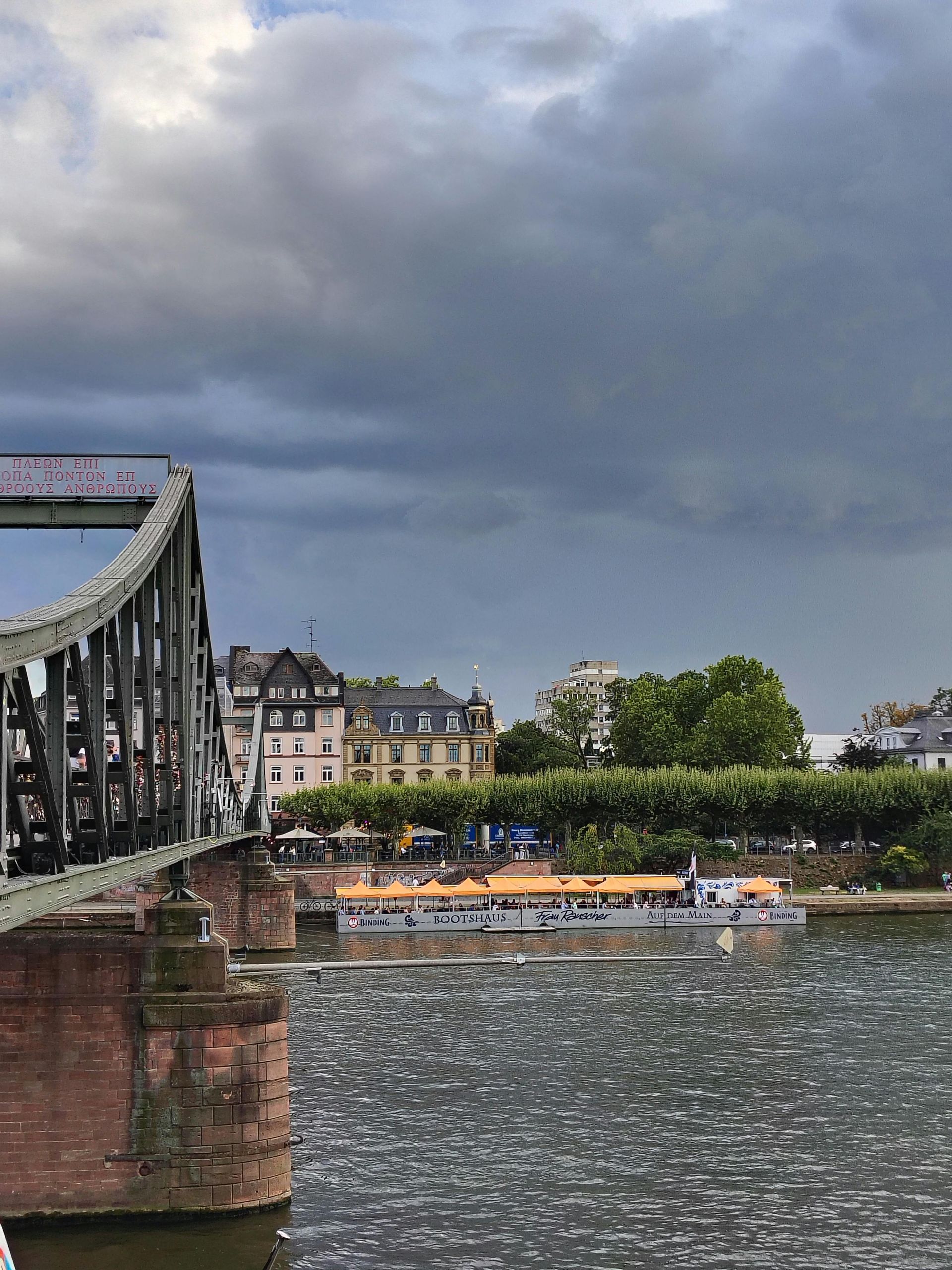 A river view with a bridge, boat, and buildings under a cloudy sky in Frankfurt, Germany.