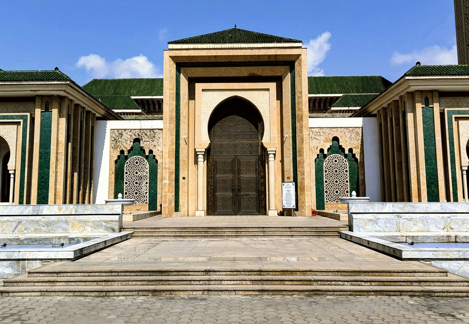 Mosque exterior with green tiled roof, arched doorway, and symmetrical design against a blue sky.