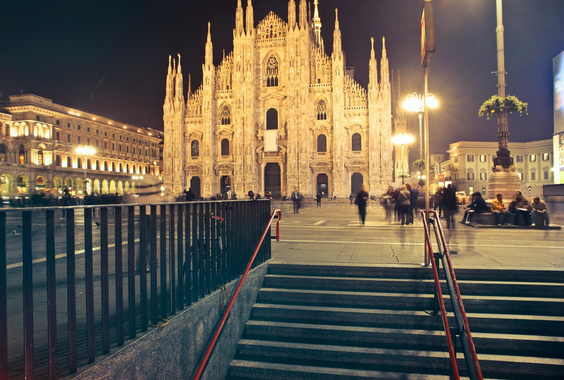 Night view of Milan Cathedral with blurred figures in the plaza, illuminated by lights; a staircase in the foreground.