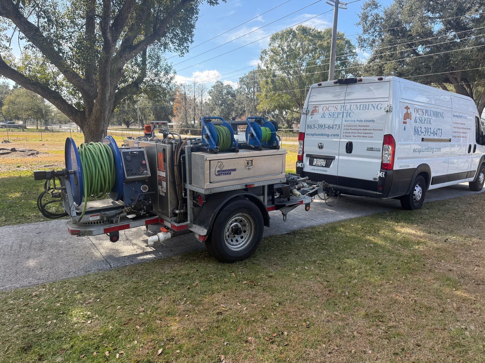 White van towing a trailer with a large hose reel on a gravel path near a tree.