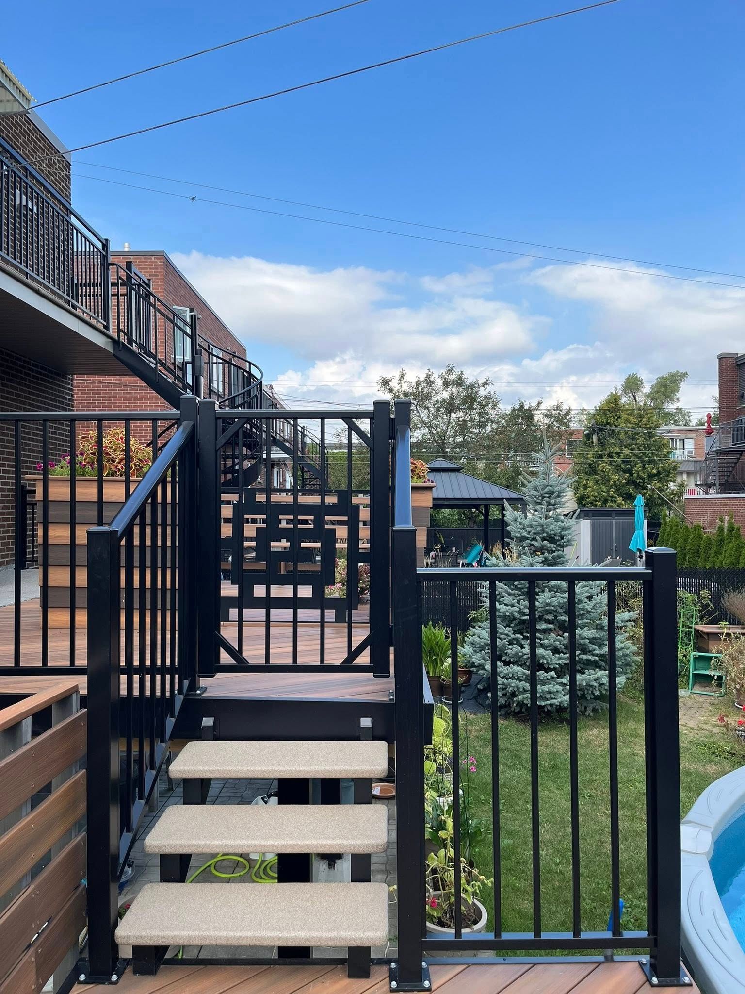 Un escalier et une rambarde en métal noir mènent à une terrasse en bois avec une piscine et un jardin verdoyant par une journée ensoleillée.