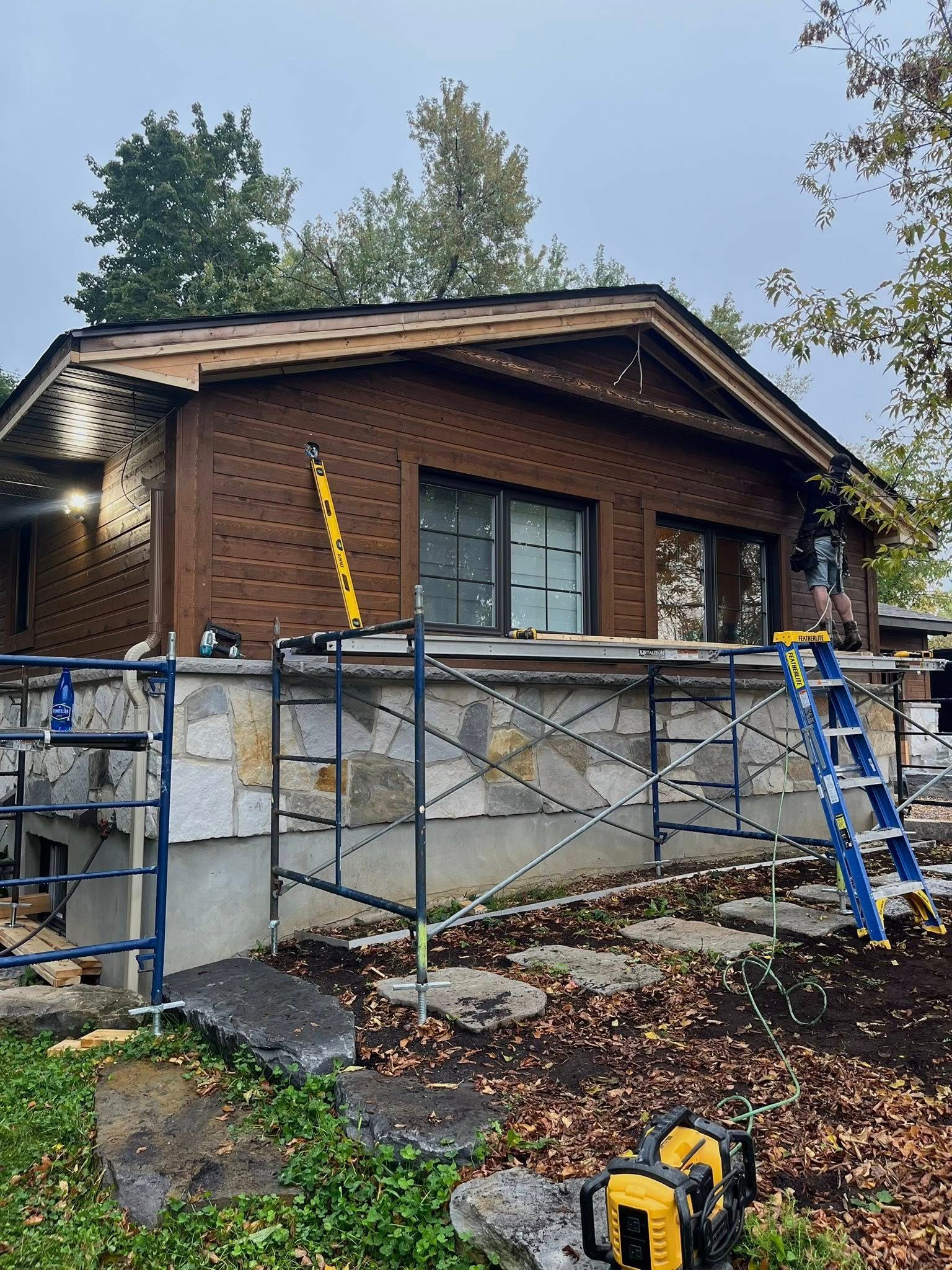 Maison en construction : bardage marron, échafaudage, échelle, charpente apparente, ciel bleu-gris, feuilles mortes.