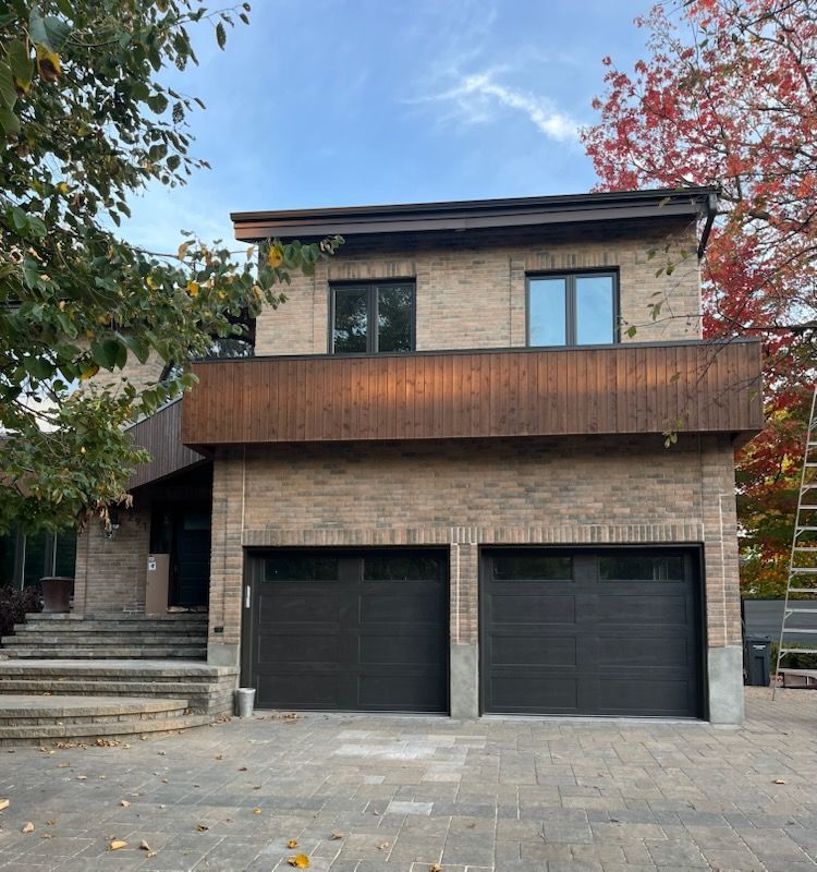 Maison en briques de deux étages avec portes de garage sombres, balcon marron, marches et arbres d'automne sur fond de ciel bleu.