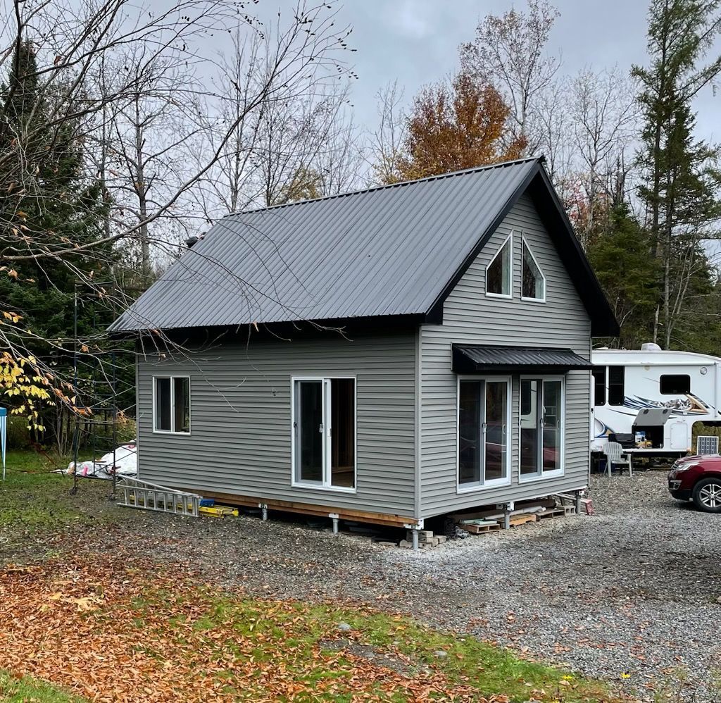 Petite cabane grise avec toit en métal, fenêtres et portes vitrées, entourée de gravier et d'arbres.