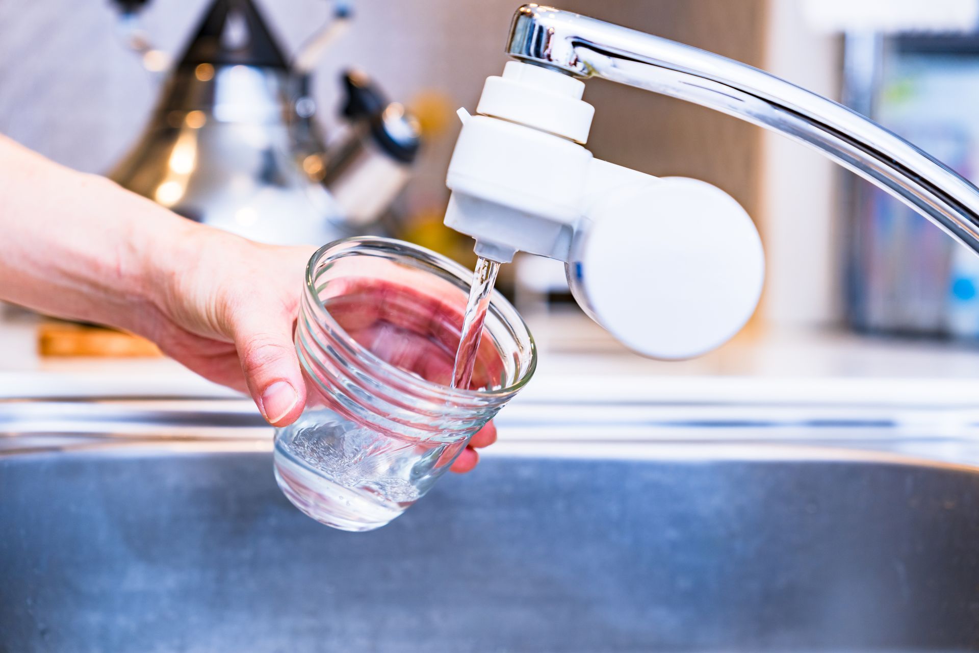A person filling a glass with water from a kitchen faucet over a stainless steel sink.