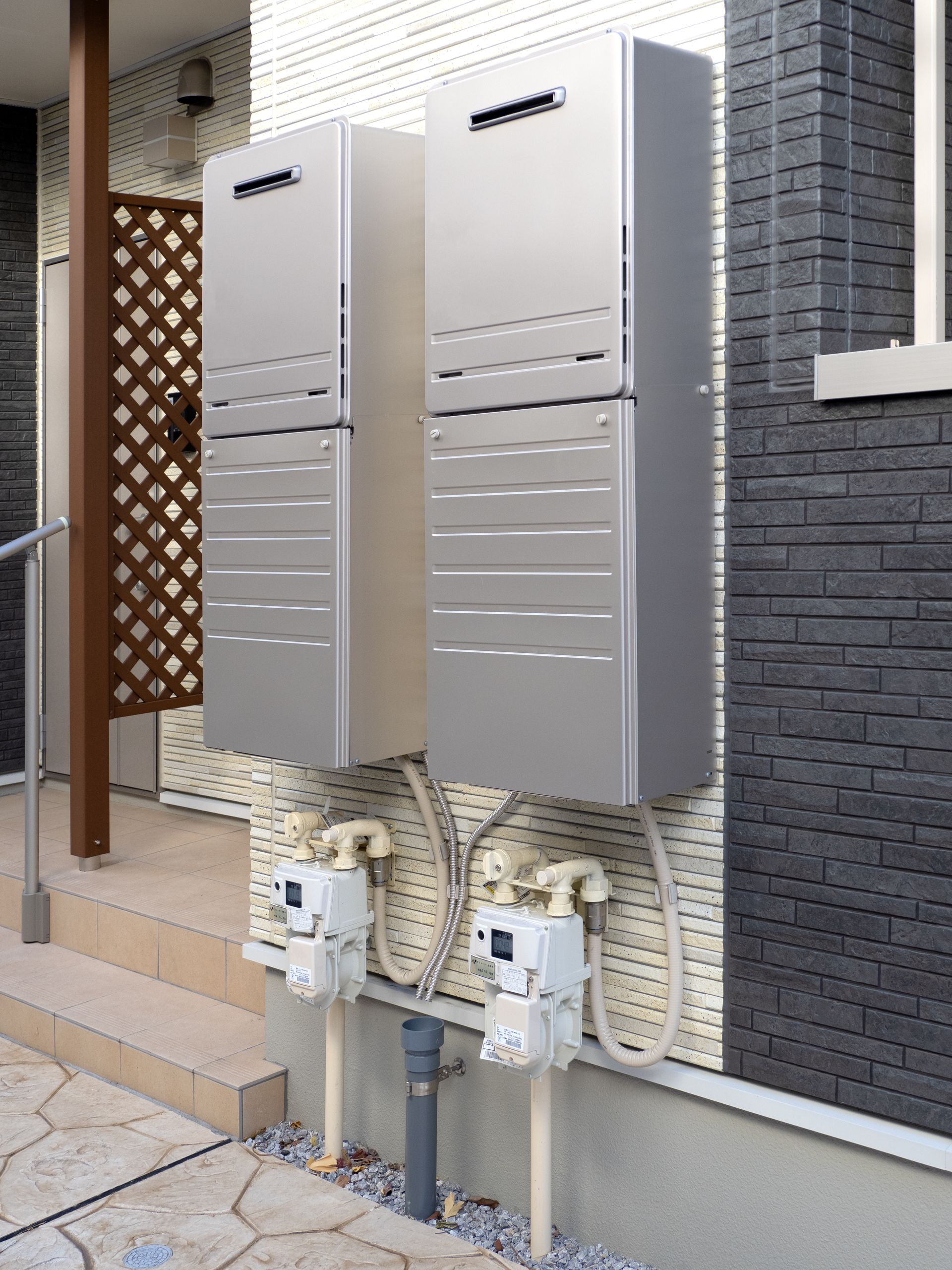 Two silver, rectangular outdoor gas water heaters mounted on a textured gray and white wall.