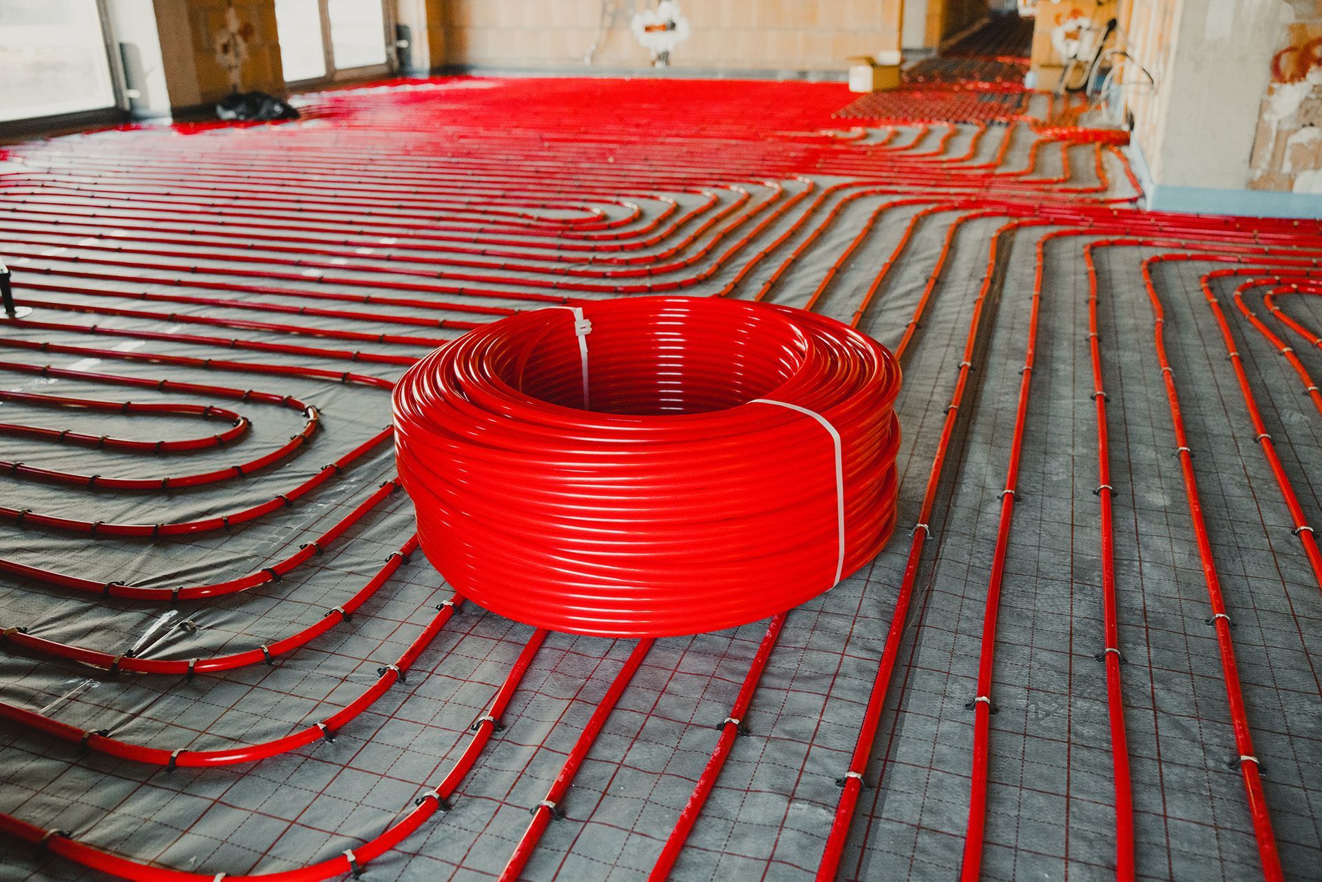 Red tubing laid on a floor, likely for radiant floor heating installation. A large coil of tubing is centered.