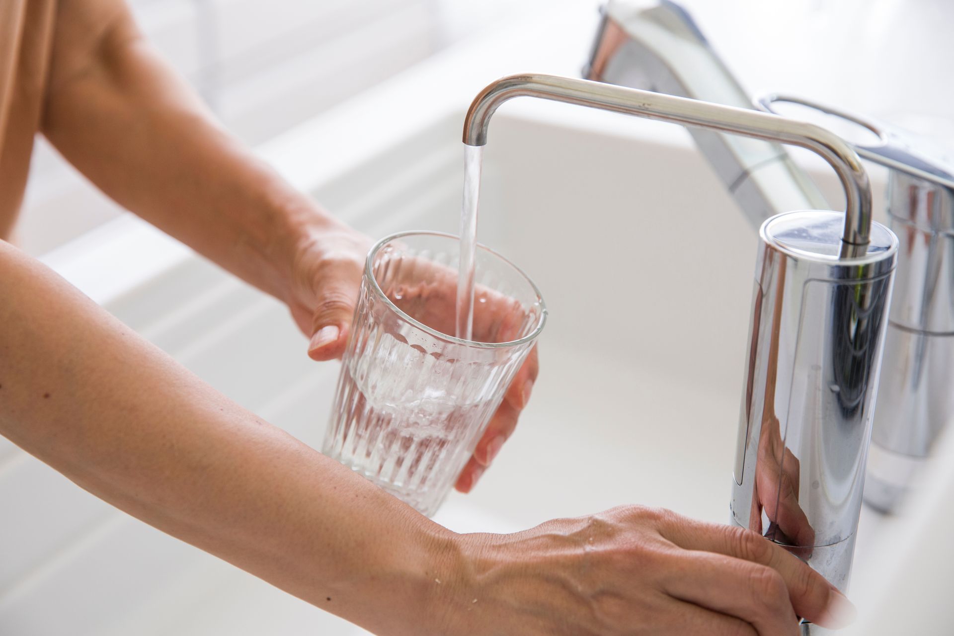 Person filling a clear glass with water from a chrome faucet in a white sink.