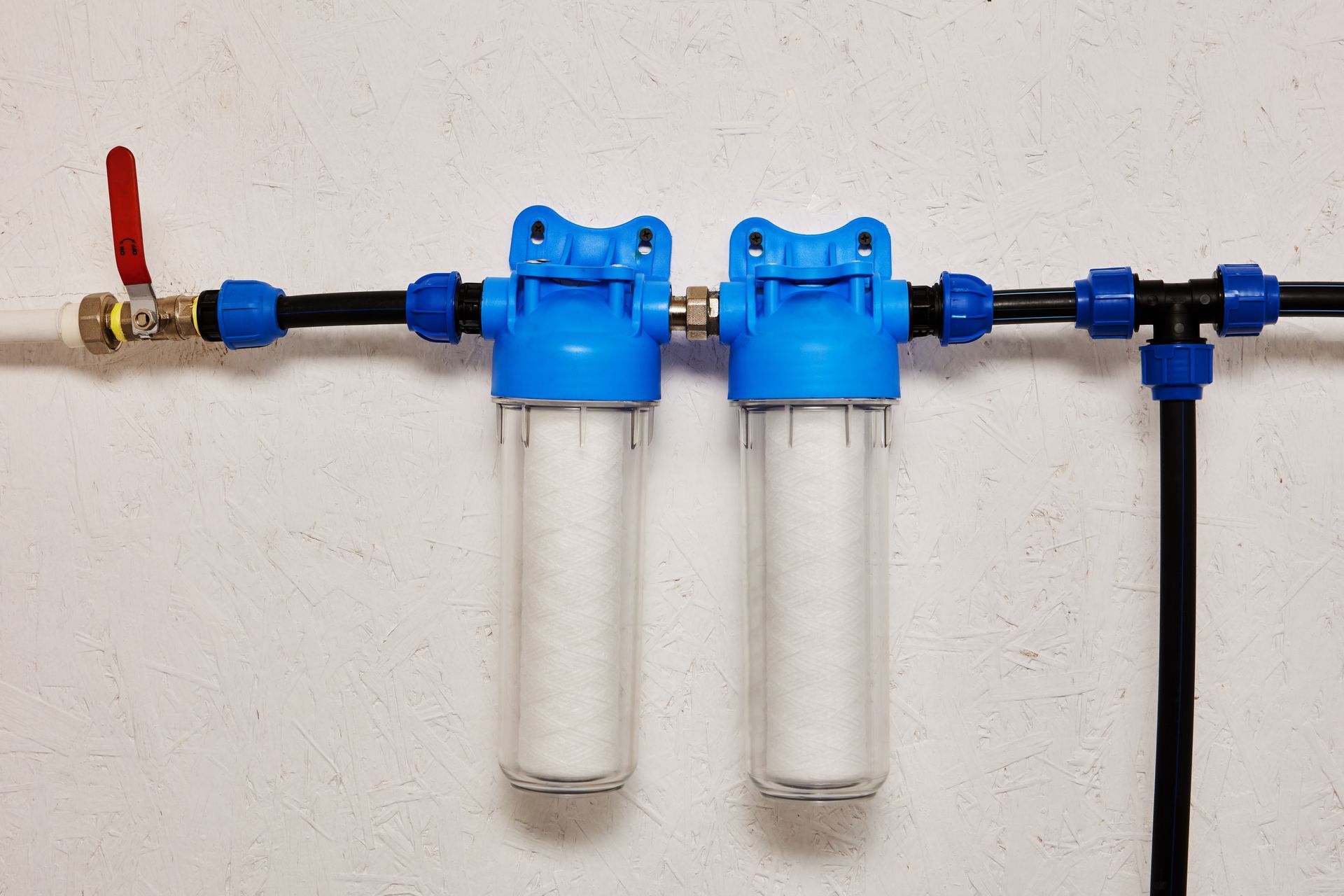 Water filter system with blue housings, clear canisters, and black pipes on a white wall.