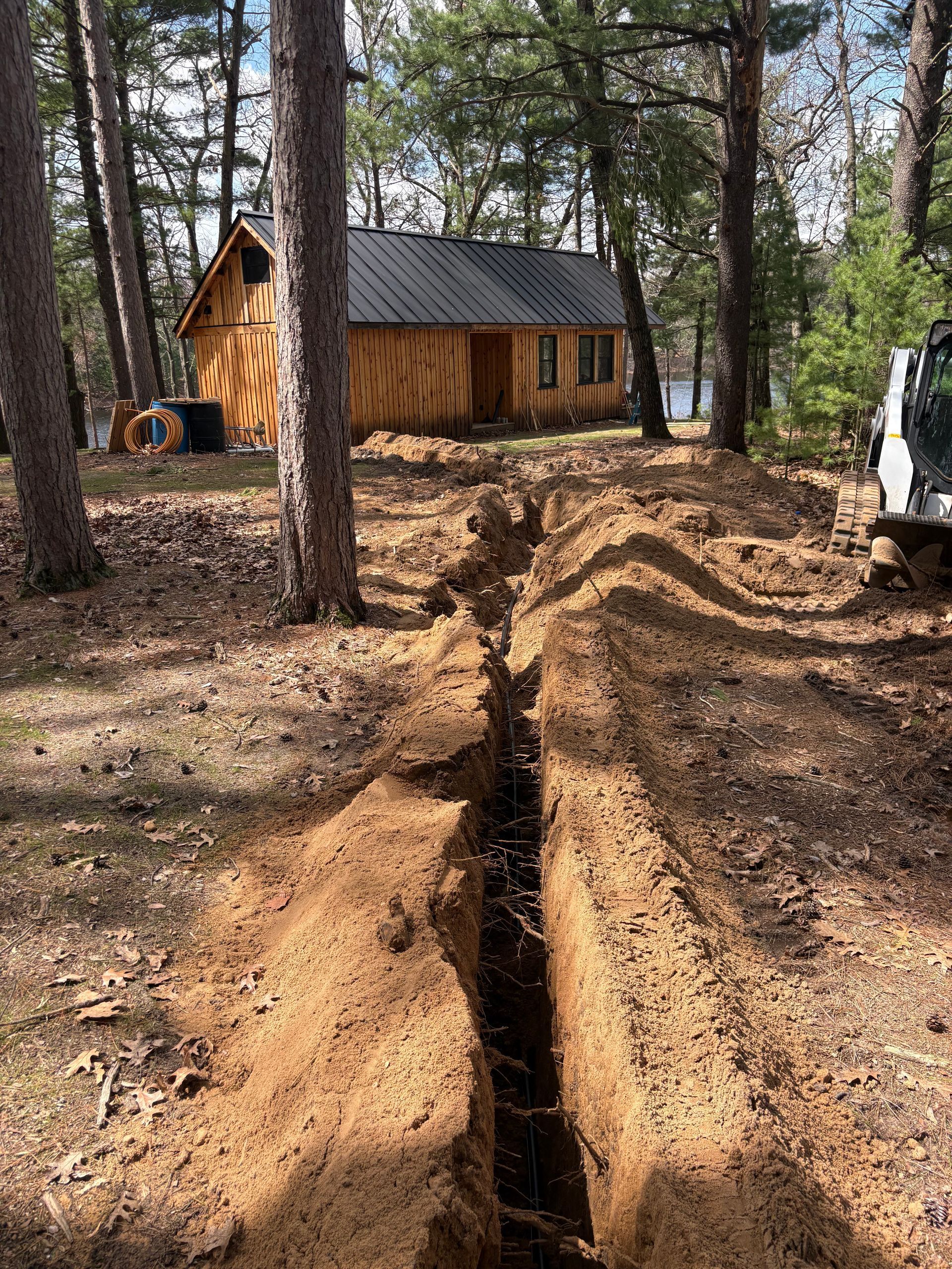 Deep trench cut through a pine forest clearing near a small wooden cabin and excavator