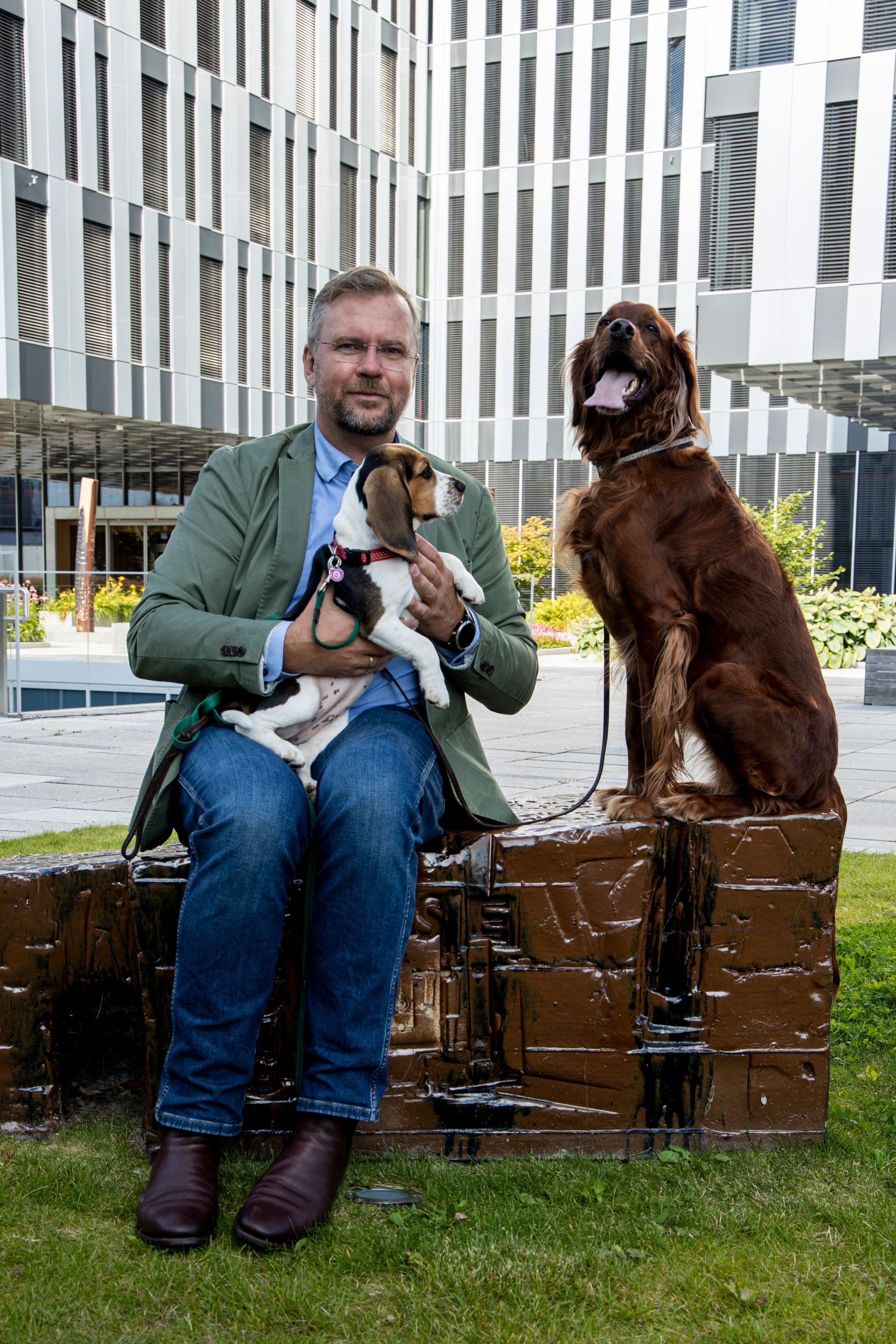 Man holding a puppy, sits with a large brown dog on a brick structure outside a modern building.