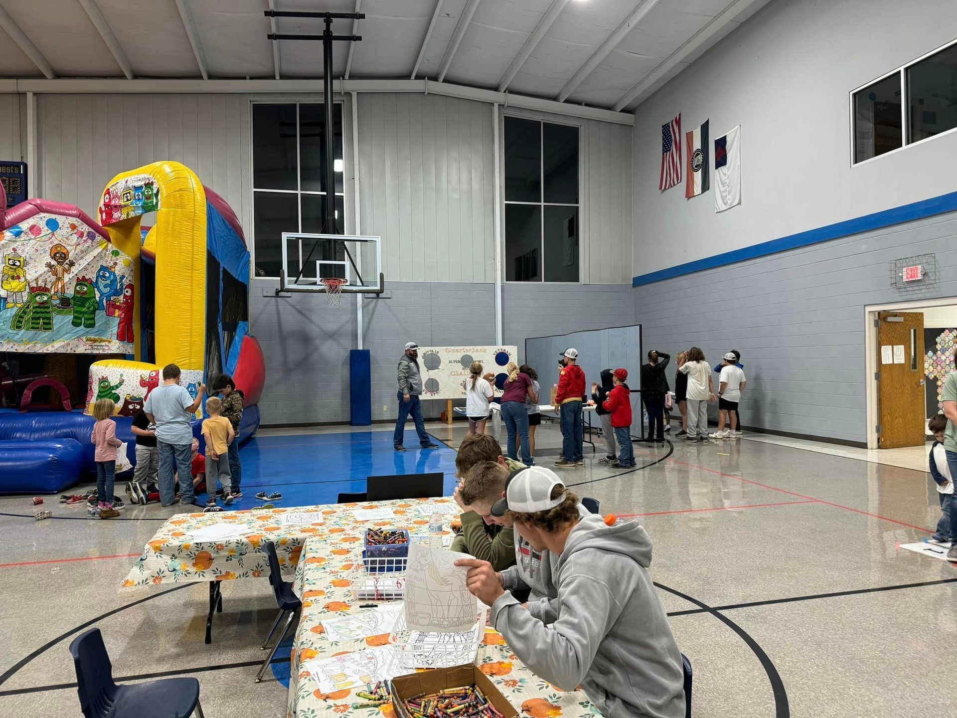 A group of people are standing around tables in a gym during Octoberfest.