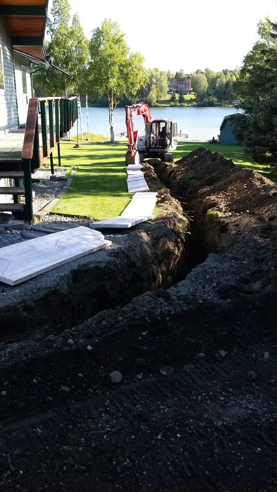 A large pile of dirt is in front of a house with a lake in the background.