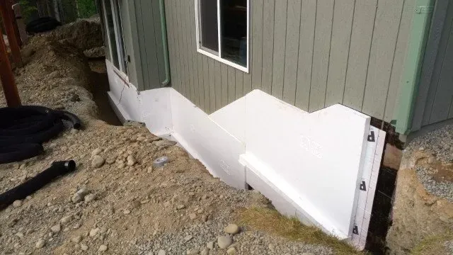 Foam insulation on a building's foundation steps down into a trench in an outdoor setting with dirt and gravel.