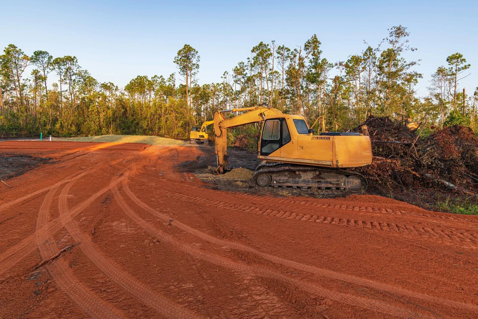 Yellow excavator on red dirt road near trees, clearing land.