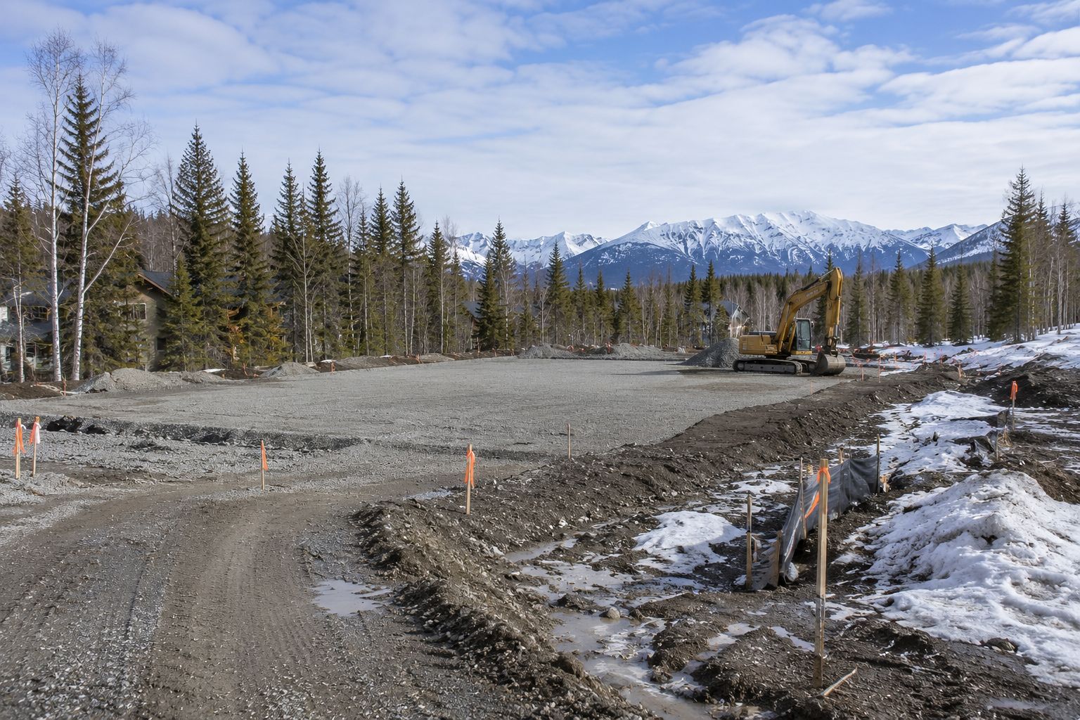 Gravel road under construction with snow patches, evergreen trees, and mountains in the distance