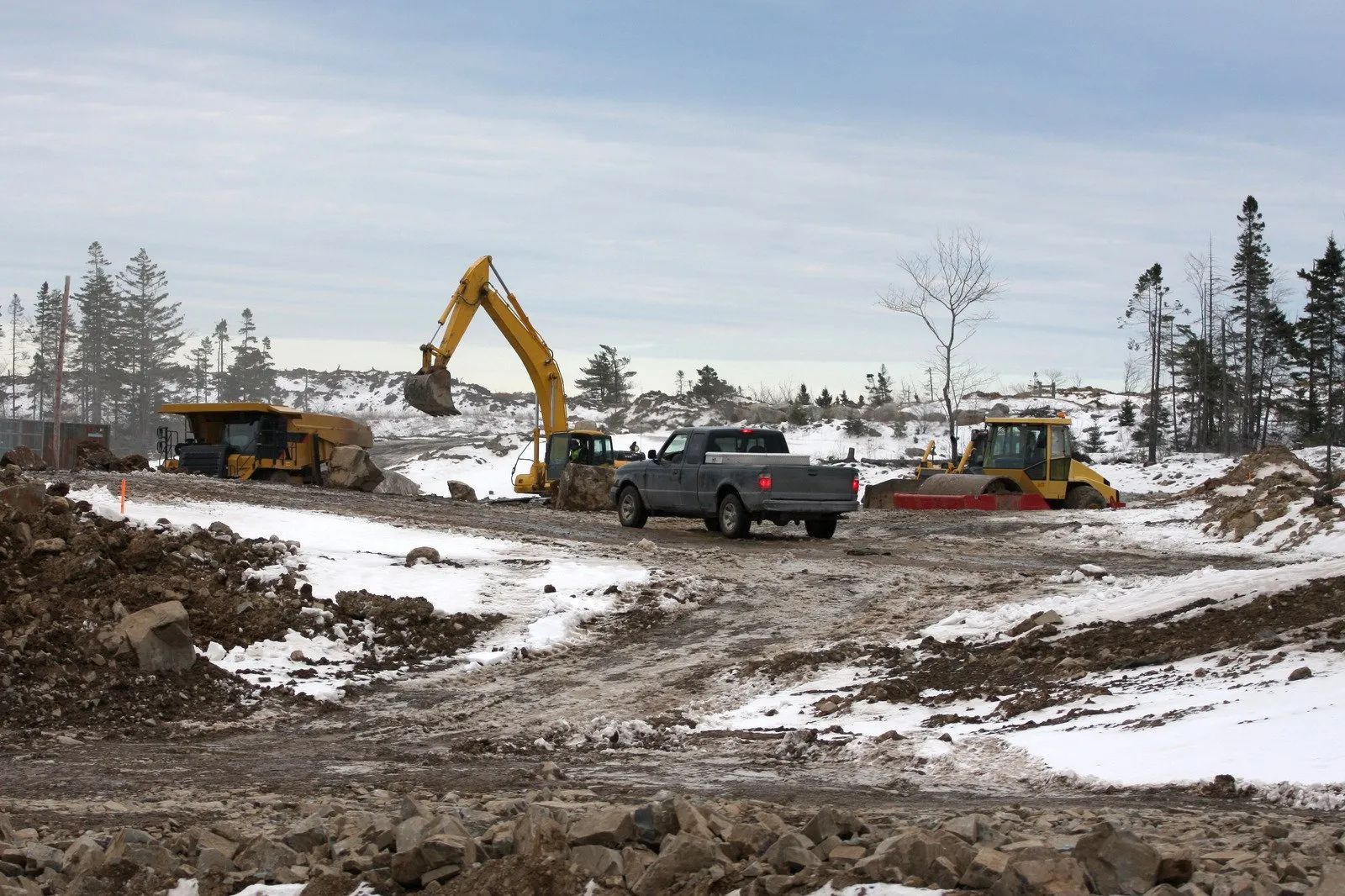 Construction site with excavator, dump truck, and snowy muddy ground under overcast sky