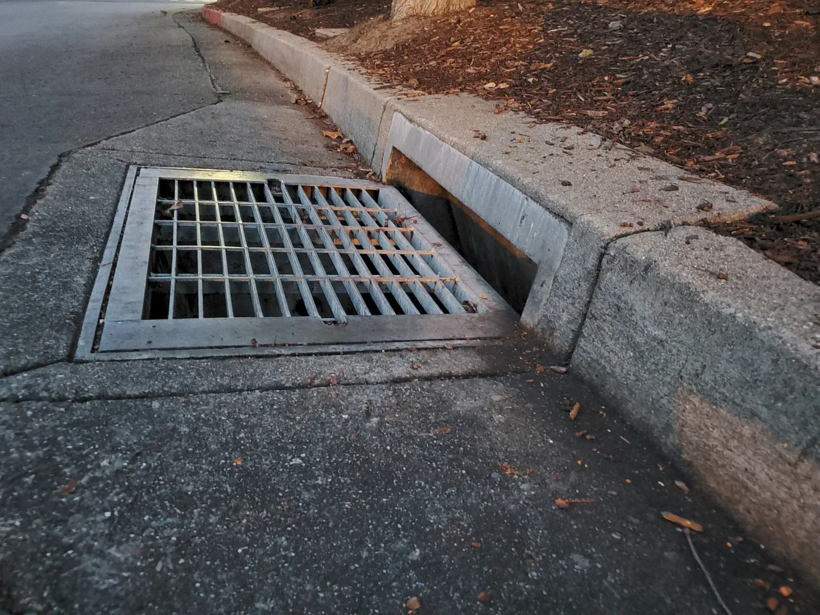 Metal storm drain in concrete curb next to asphalt road and dirt.