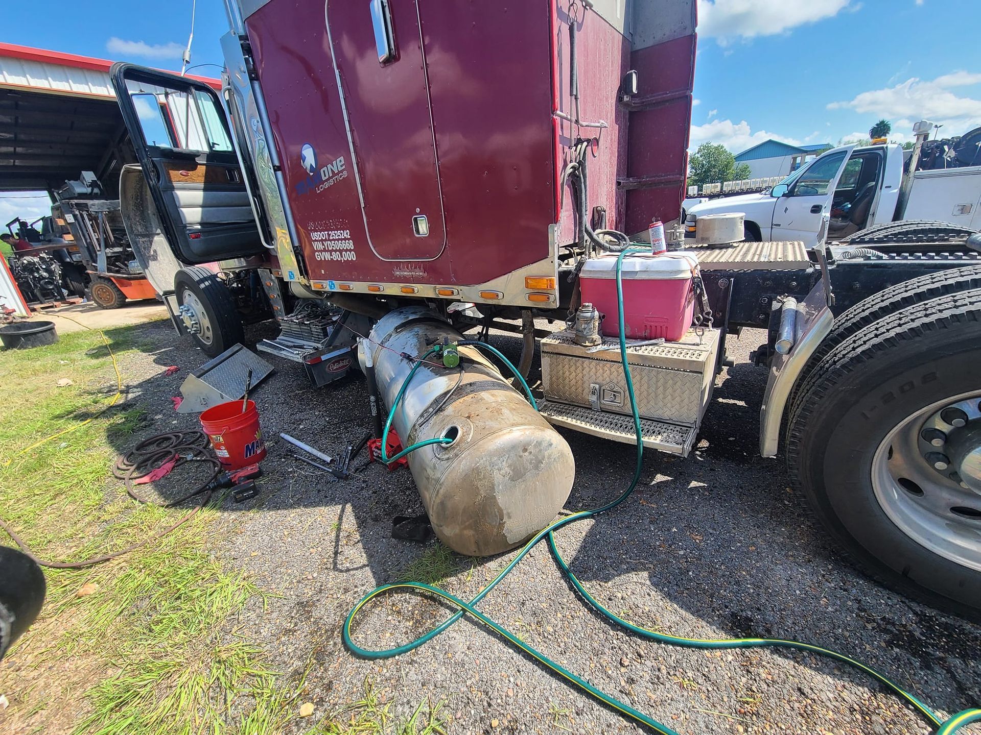 A burgundy semi-truck with a detached fuel tank on grass. A cooler sits on the truck frame.