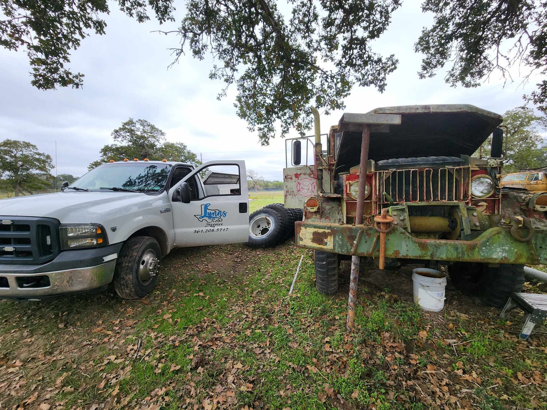 White pickup truck next to a rusty, vintage green military truck, both parked outdoors under trees.