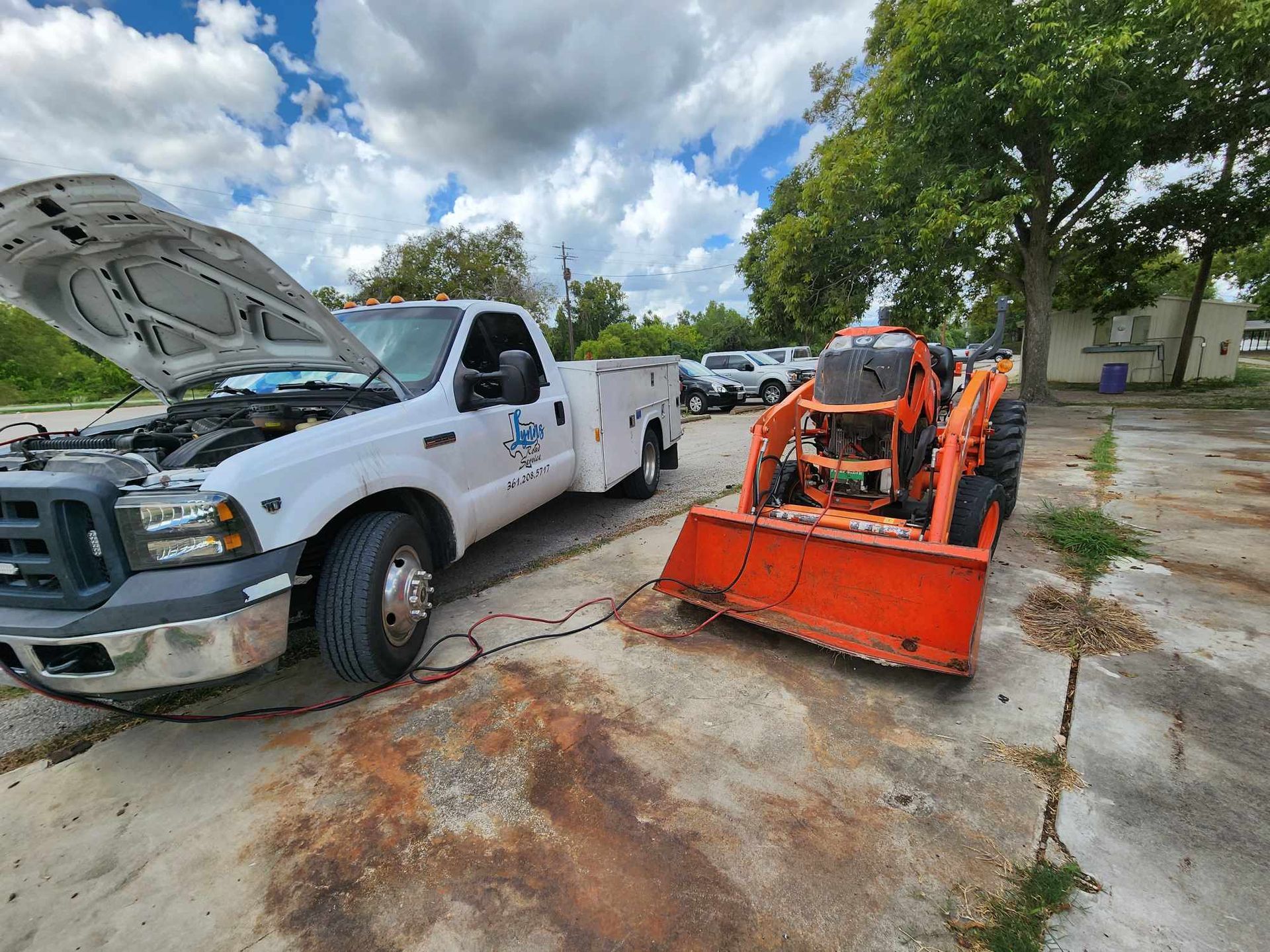 White truck jump-starting orange skid steer in a paved lot under a cloudy sky.