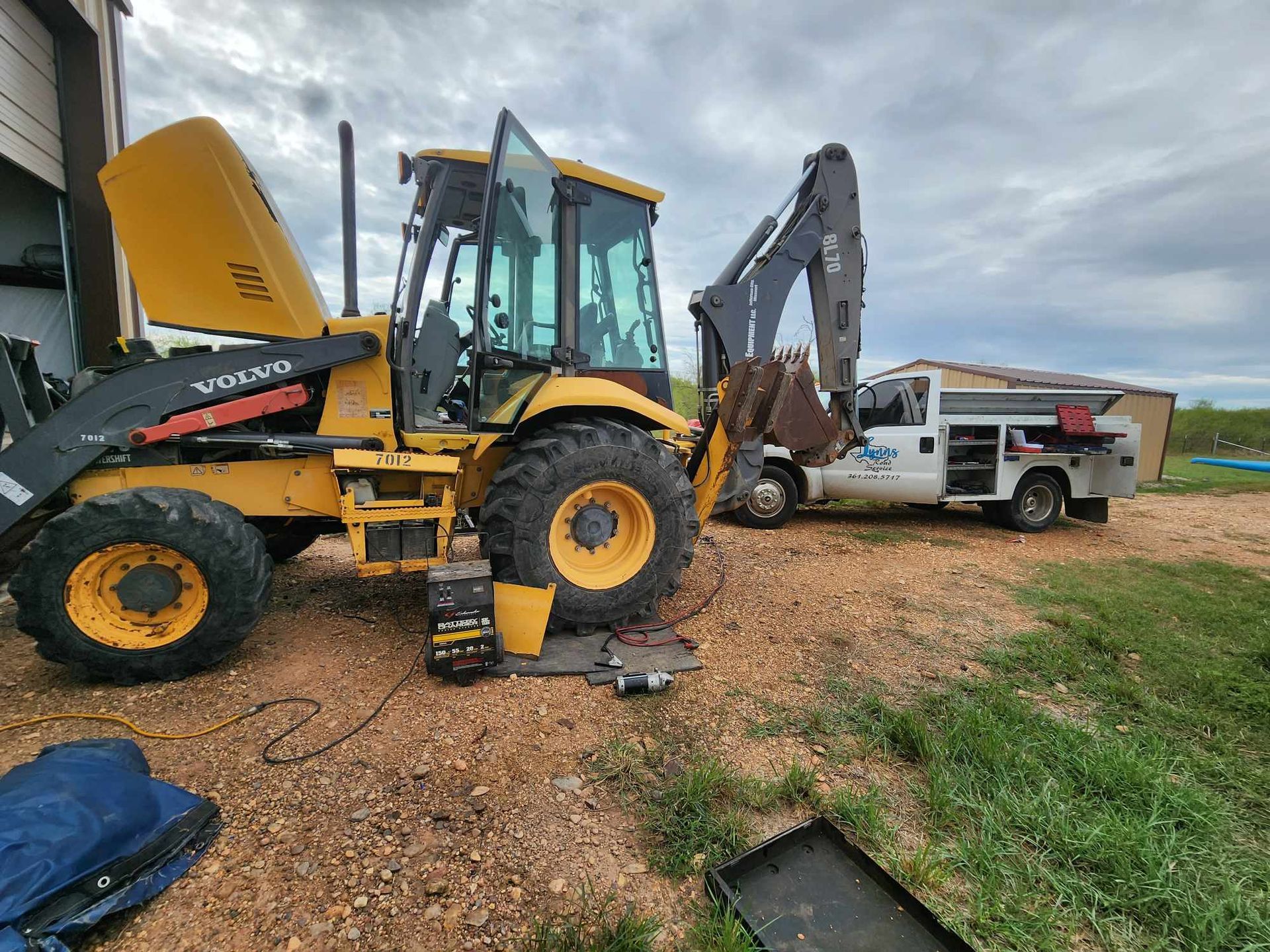 Yellow backhoe with its hood up, parked near a service truck, outdoors.