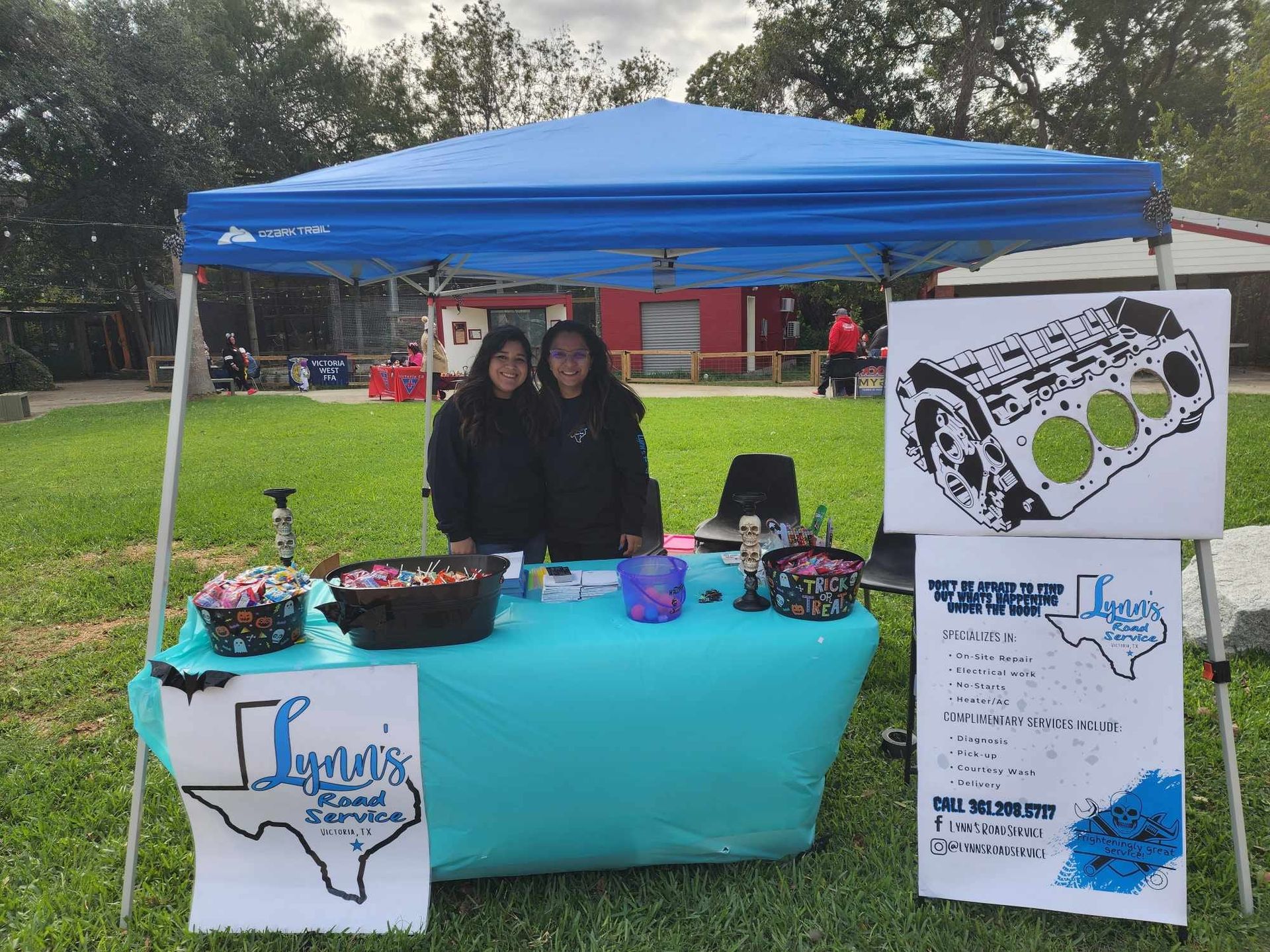Two women at a booth, selling auto parts. Blue canopy, teal table. Texas-themed logo, grass background.