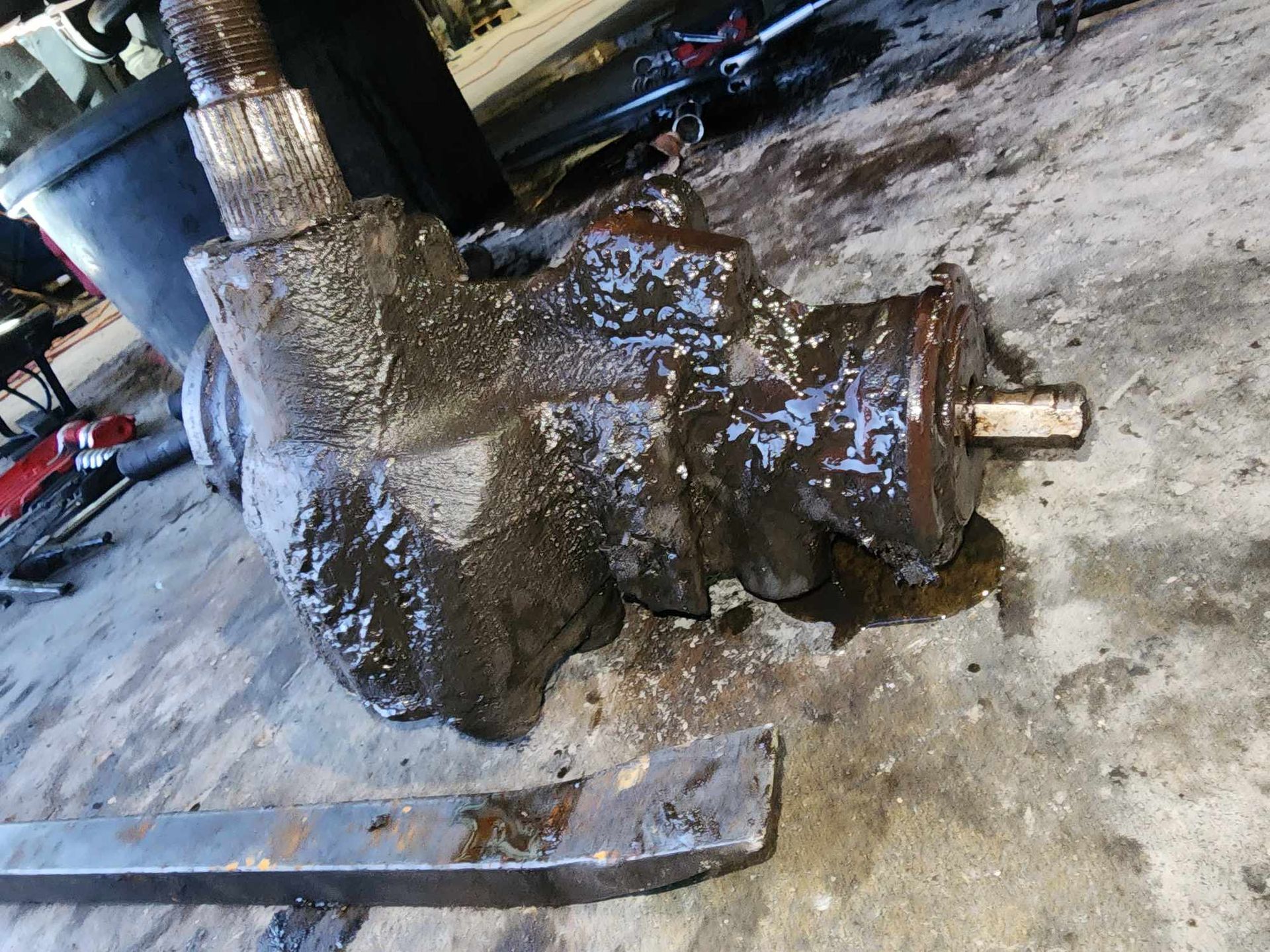 A greasy, rusty steering gear box on a concrete surface. A crowbar is in the foreground.