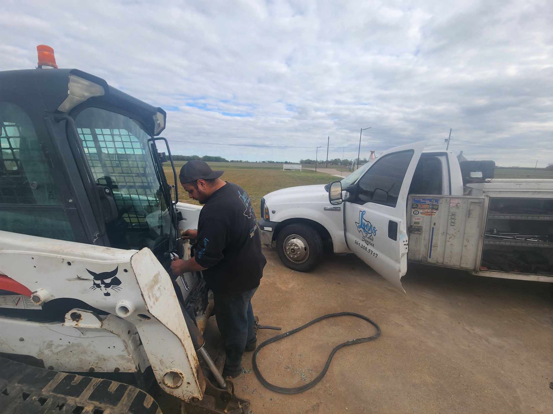 A man works on a Bobcat while a white work truck is parked nearby on a cloudy day.