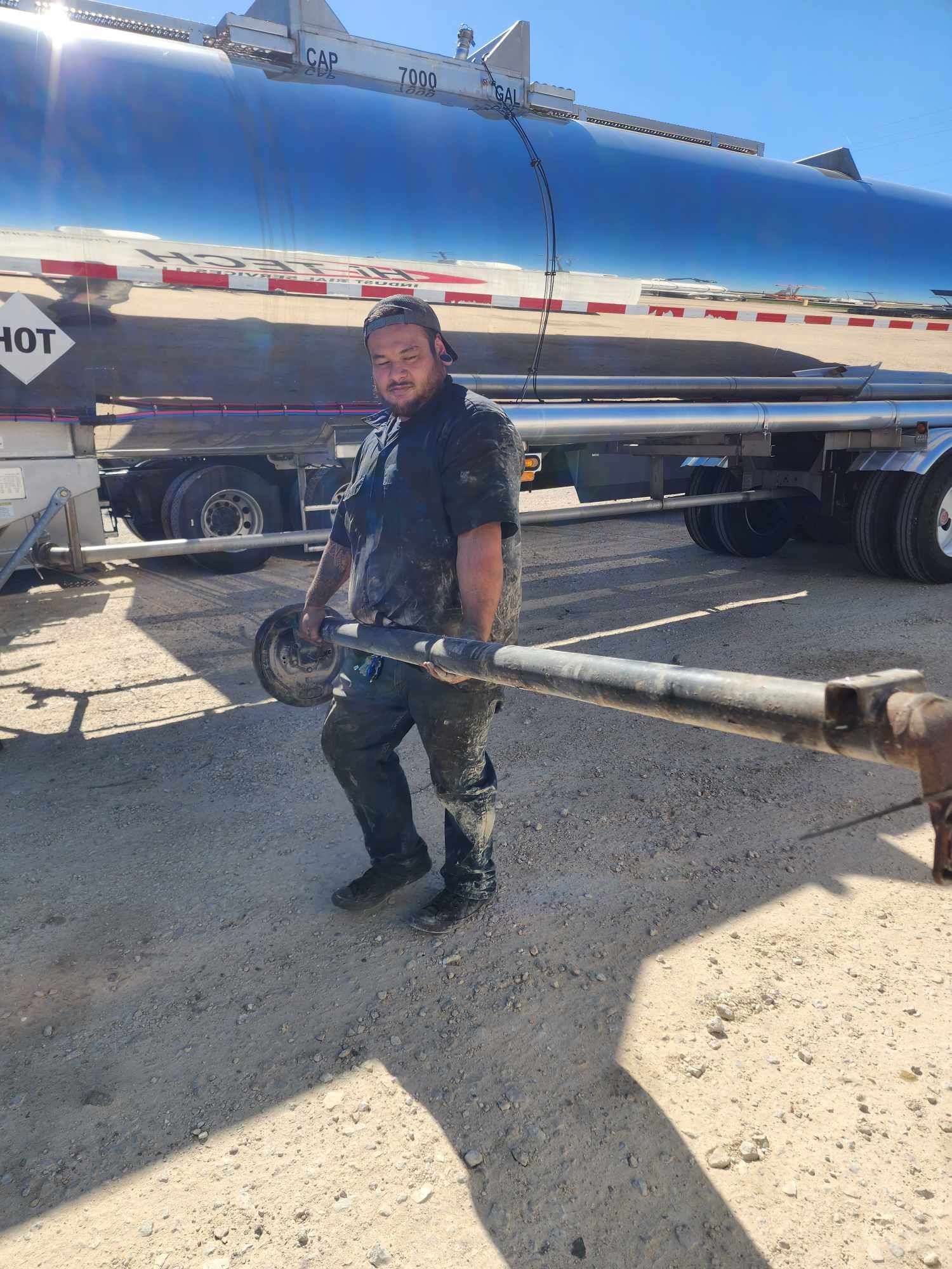 Man carrying metal pipe, standing near a tanker truck on a gravel surface in bright sunlight.