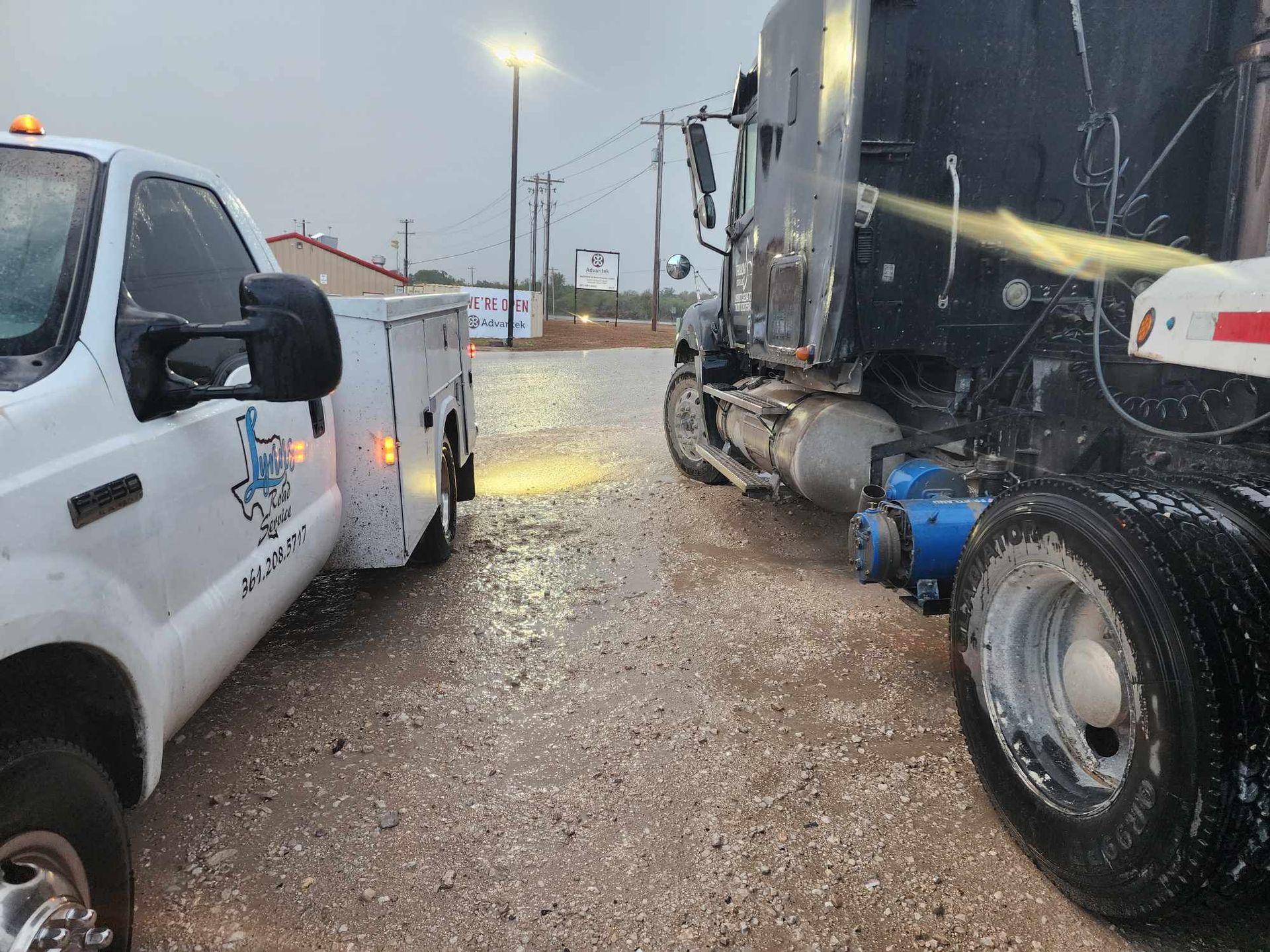 A white service truck and a black semi-truck parked on a wet road, next to a sign and under street lights.