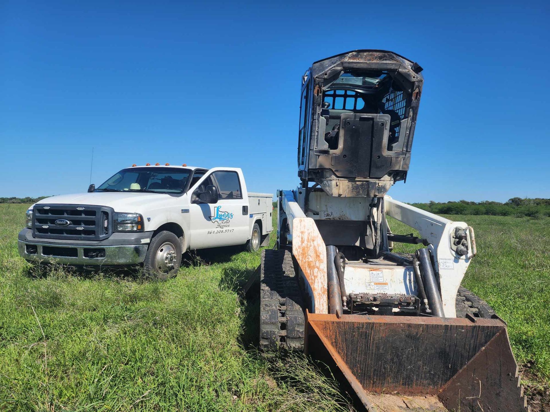 A white pickup truck and a skid steer sit in a grassy field under a blue sky.